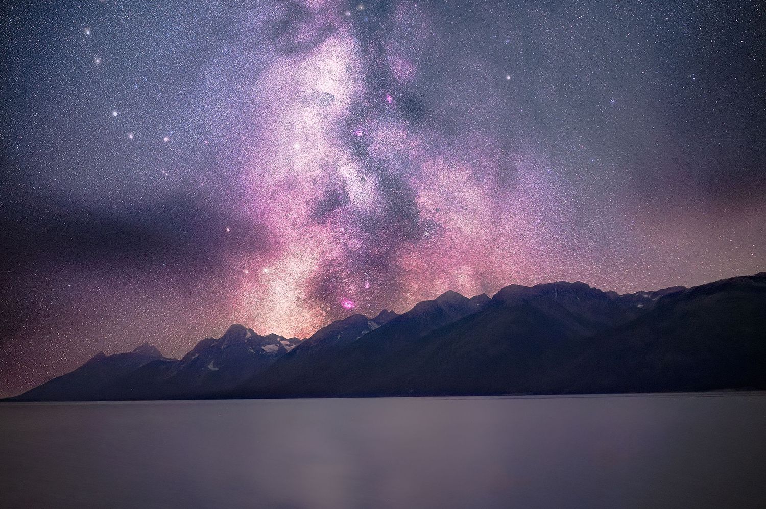 Purple and pink Milky Way galaxy stretches across mountain silhouettes at night.