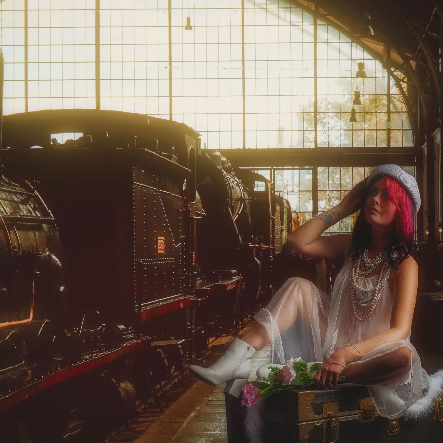 Woman with pink hair in white dress and hat poses on vintage trunk beside steam locomotives in a sunlit train depot.