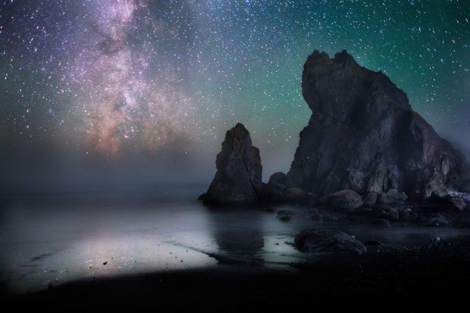 Milky Way galaxy glowing over dramatic rocky sea stacks on a misty coastline under a star-filled night sky.