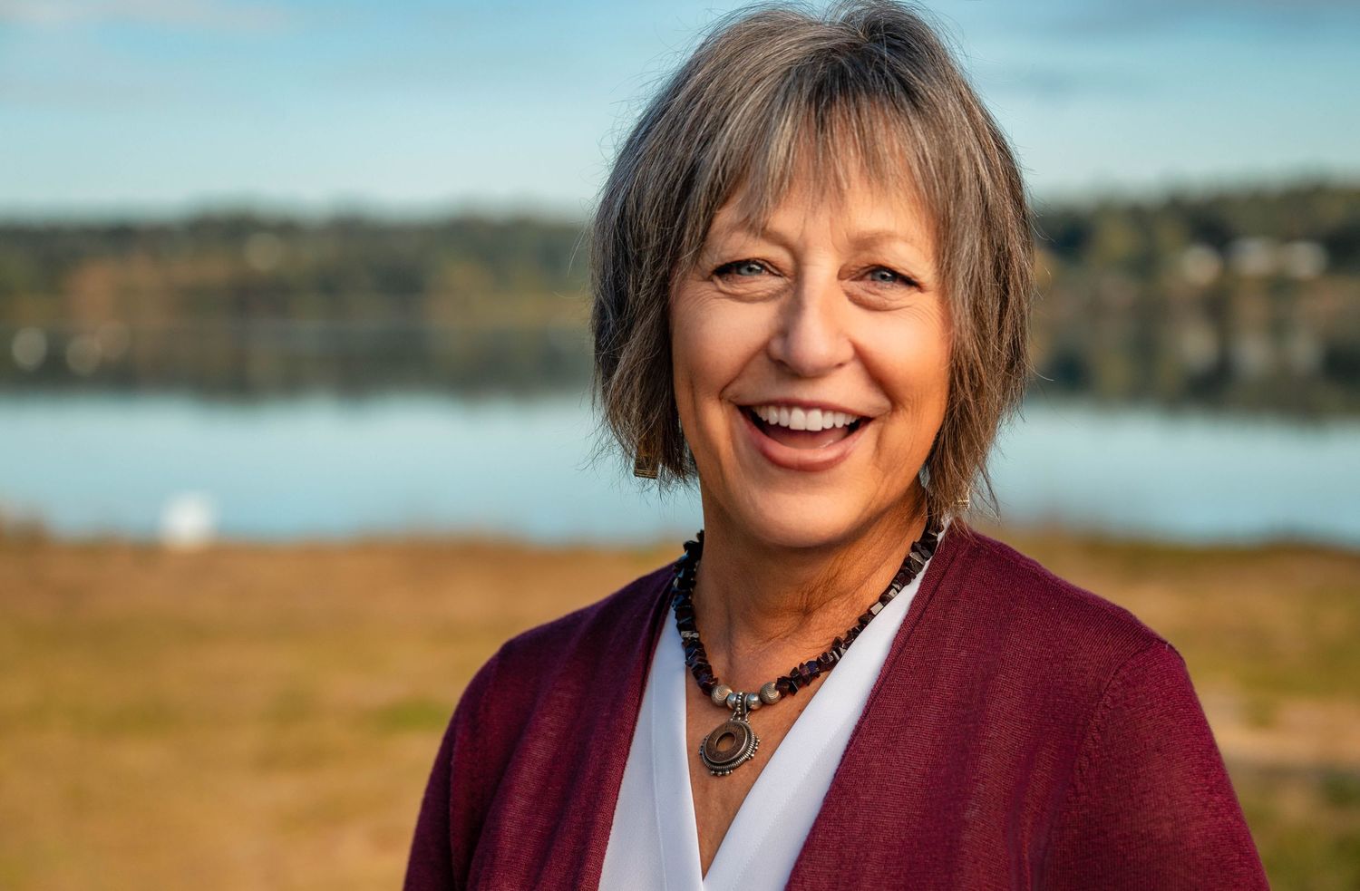 A person in a burgundy sweater smiles warmly while posing outdoors against a scenic autumn landscape background.