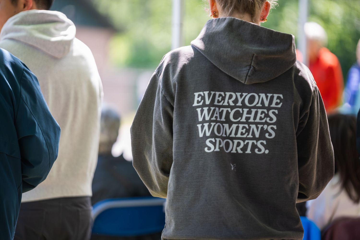 Person wearing a gray hoodie with 'Everyone Watches Women's Sports' printed on the back at an outdoor event.