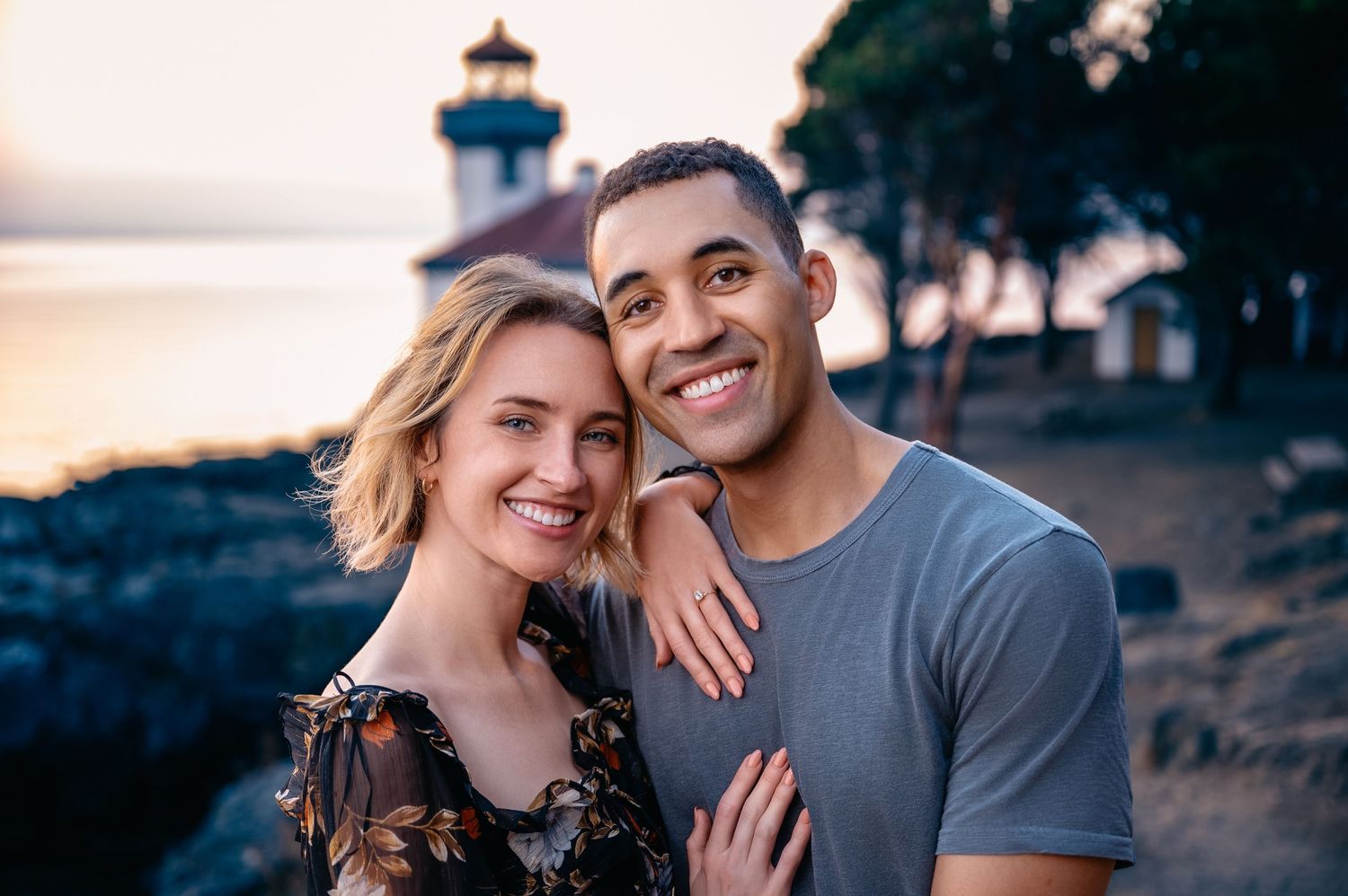 A happy couple poses together at sunset near a lighthouse on the coast.