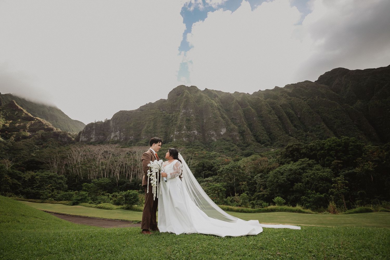 Bride and groom embrace at mountain wedding, holding white orchid bouquet with lush green cliffs and tropical landscape backdrop.