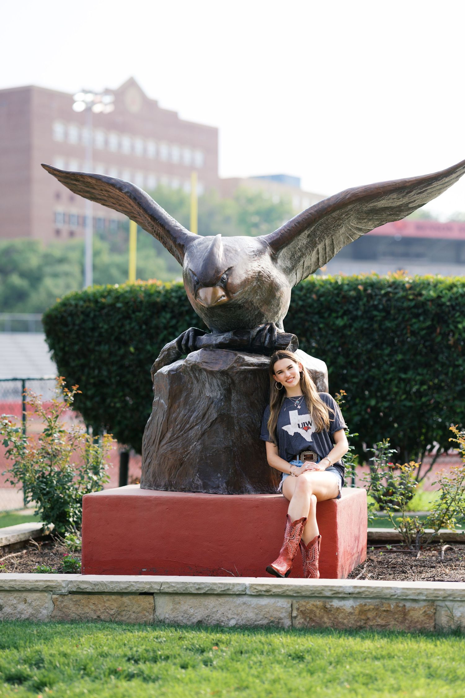 Young woman in boots poses beside a large bronze eagle statue on a red pedestal in a college campus garden.