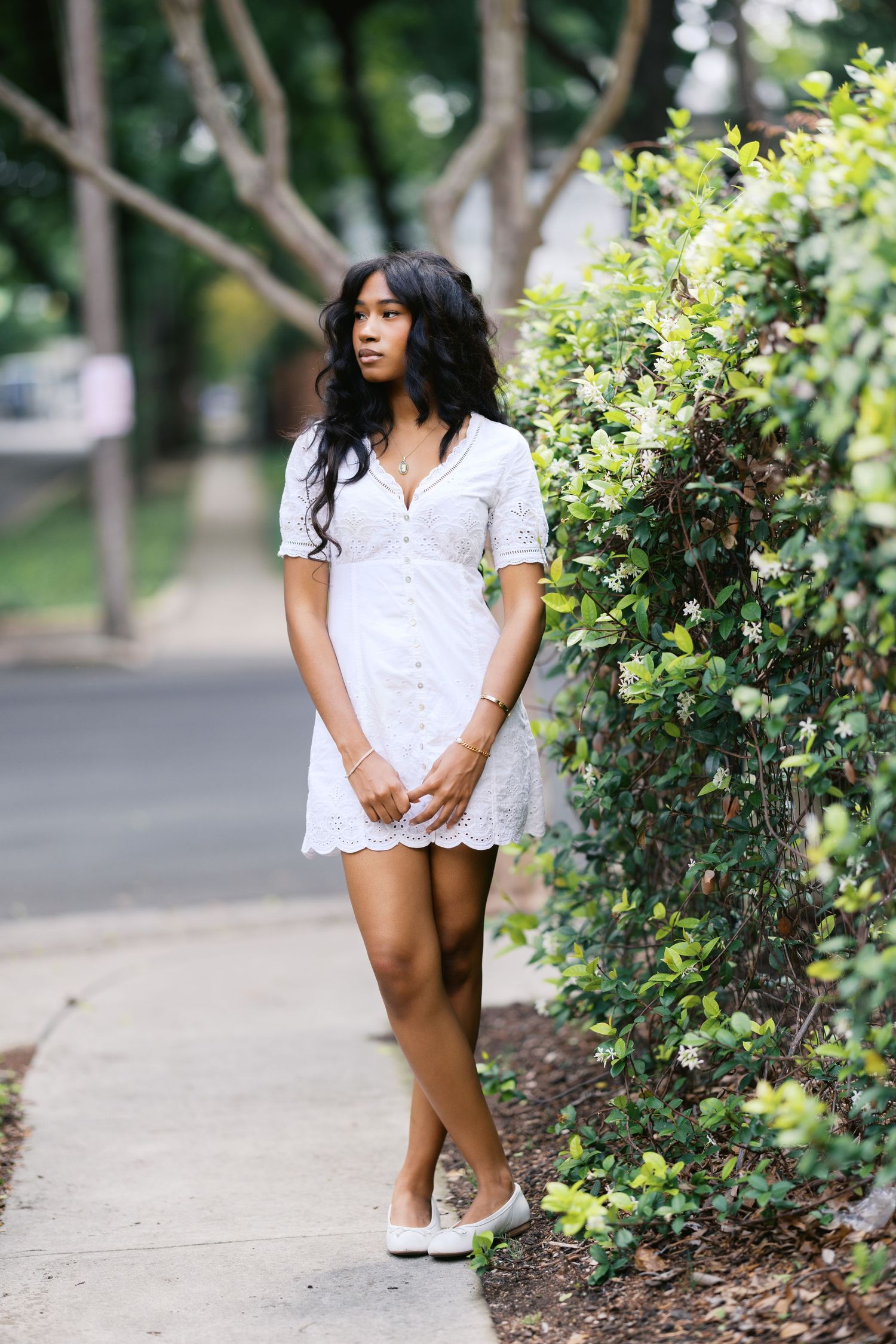 Young Black woman in white eyelet mini dress and white flats standing on sidewalk beside green hedge.