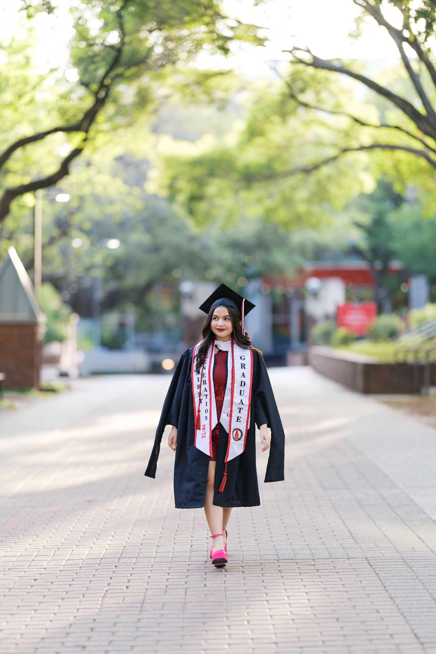 Graduate in black cap and gown with red graduation stole walks confidently on brick campus path.