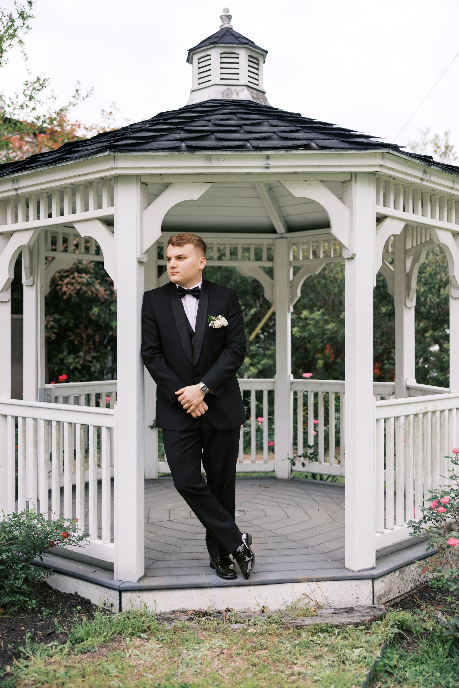 Groom in black tuxedo with bow tie and boutonniere posing in white wooden gazebo surrounded by garden greenery.