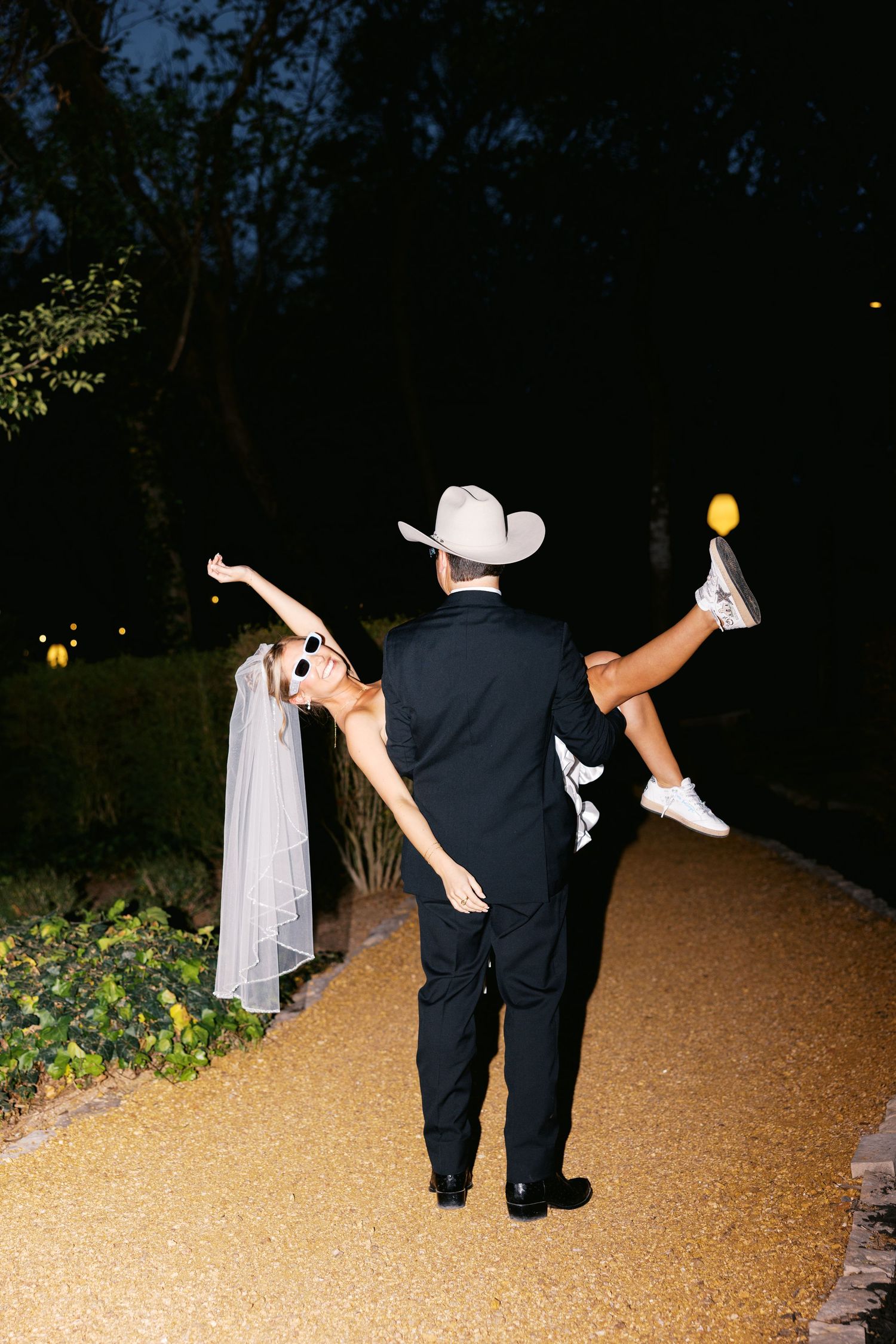 Groom in cowboy hat lifting bride in white dress and sneakers on a dark outdoor pathway at night.