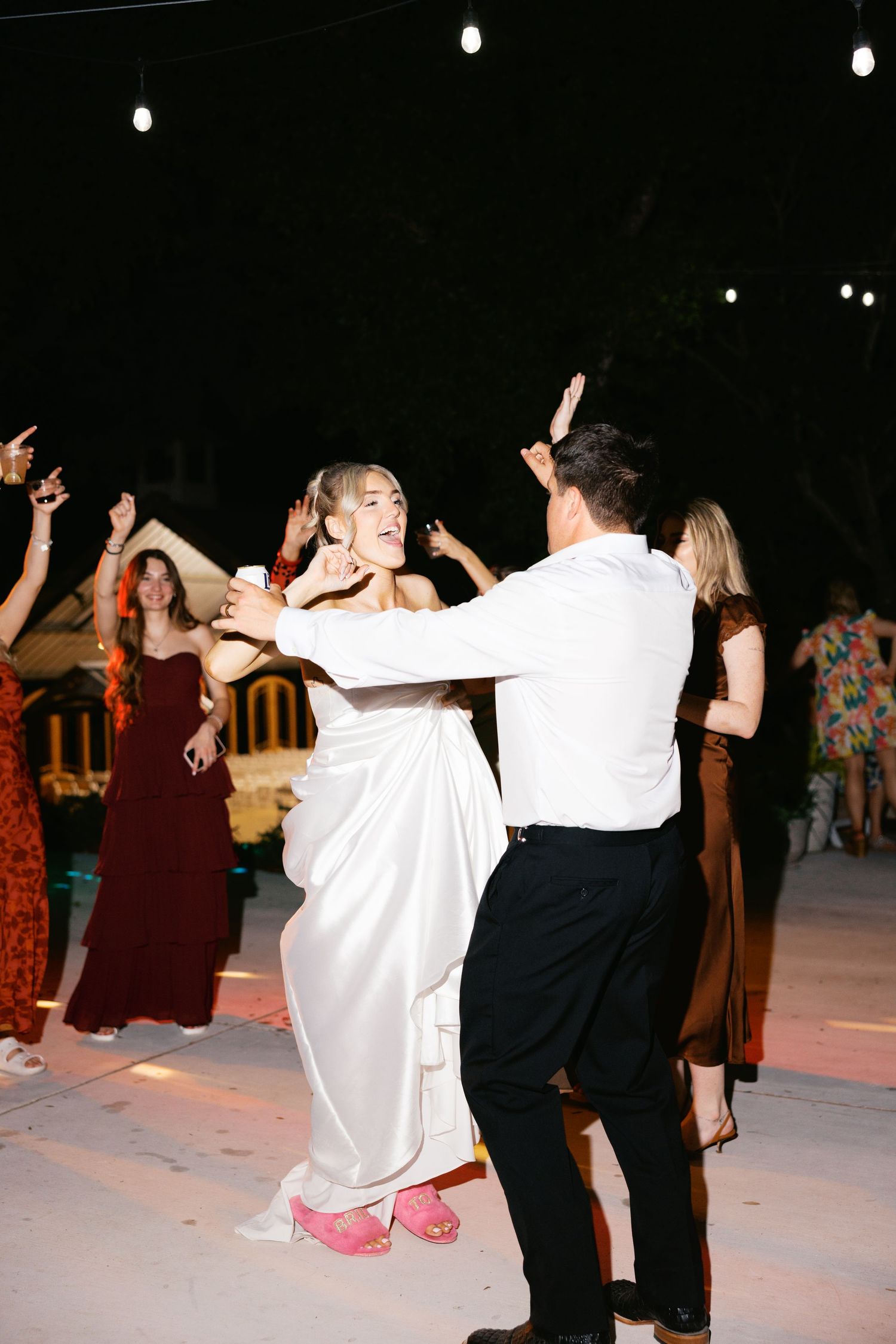Bride in white gown with pink heels dances with groom surrounded by guests at outdoor wedding reception.