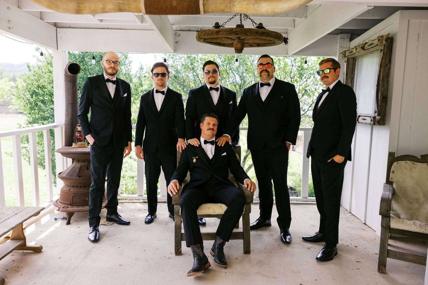 Groom seated with five groomsmen standing behind him, all wearing black tuxedos with bow ties on a rustic porch.