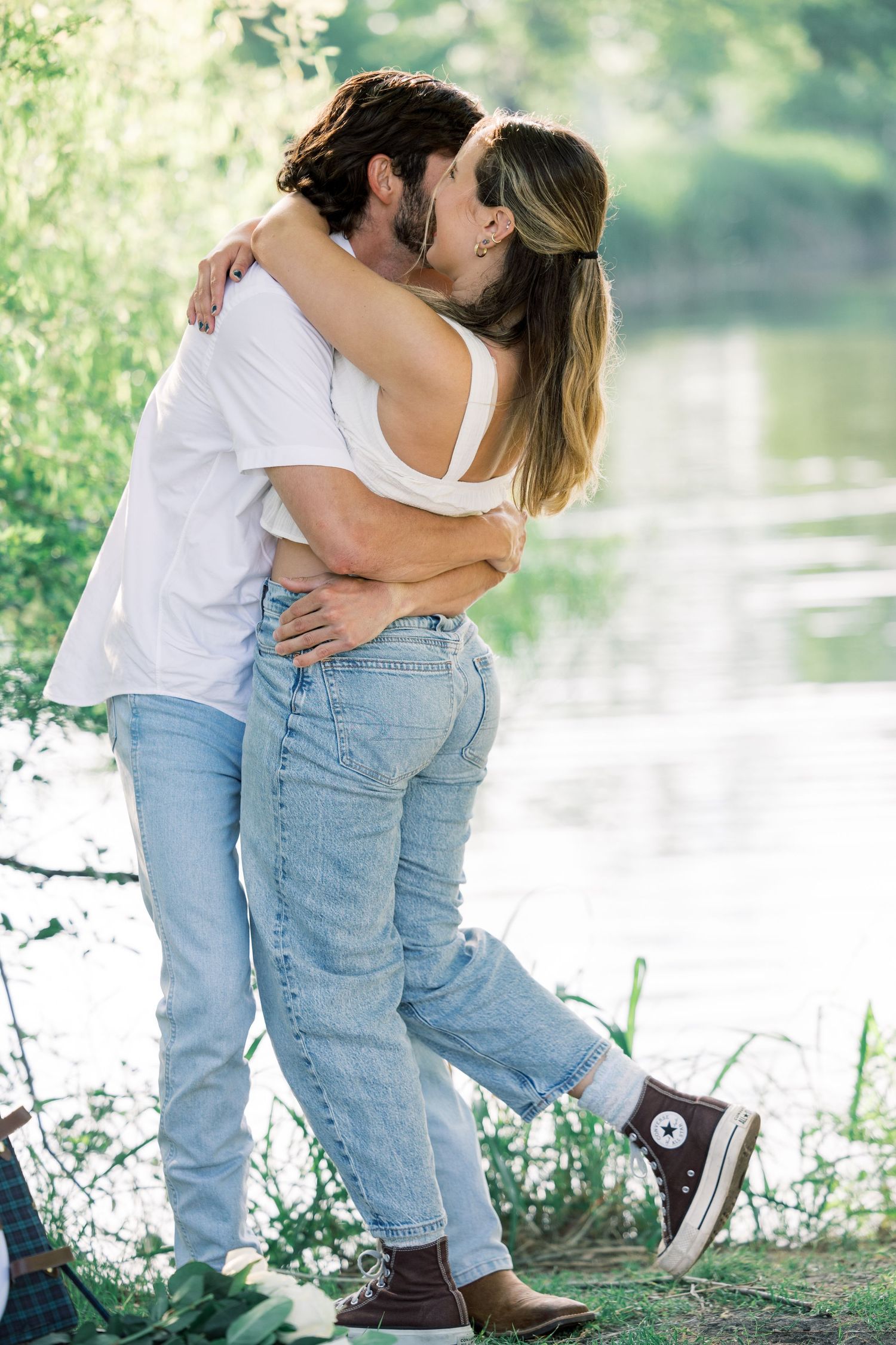 A tender embrace between two people in casual attire against a lakeside backdrop.