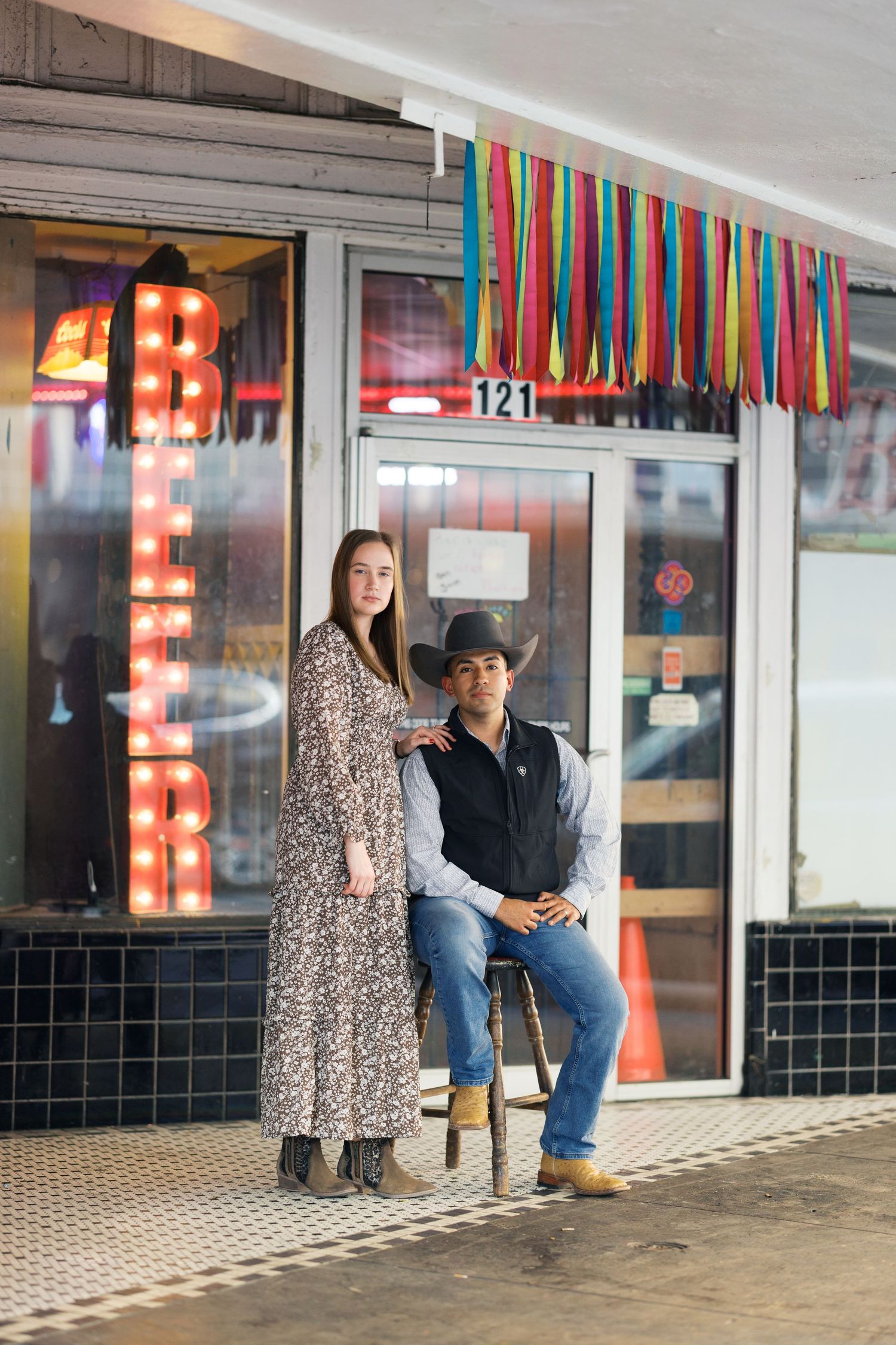 A couple posing in front of a storefront decorated with colorful streamers and lights.