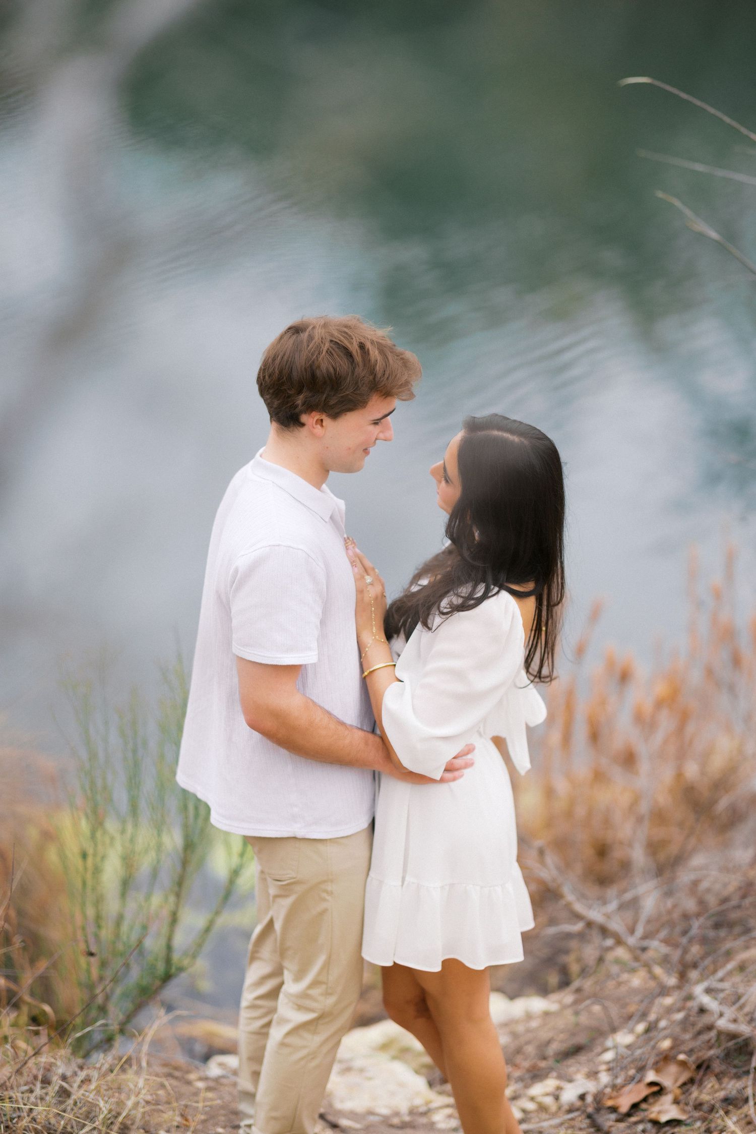 Romantic couple in white and khaki outfits share tender moments by a tranquil lake.