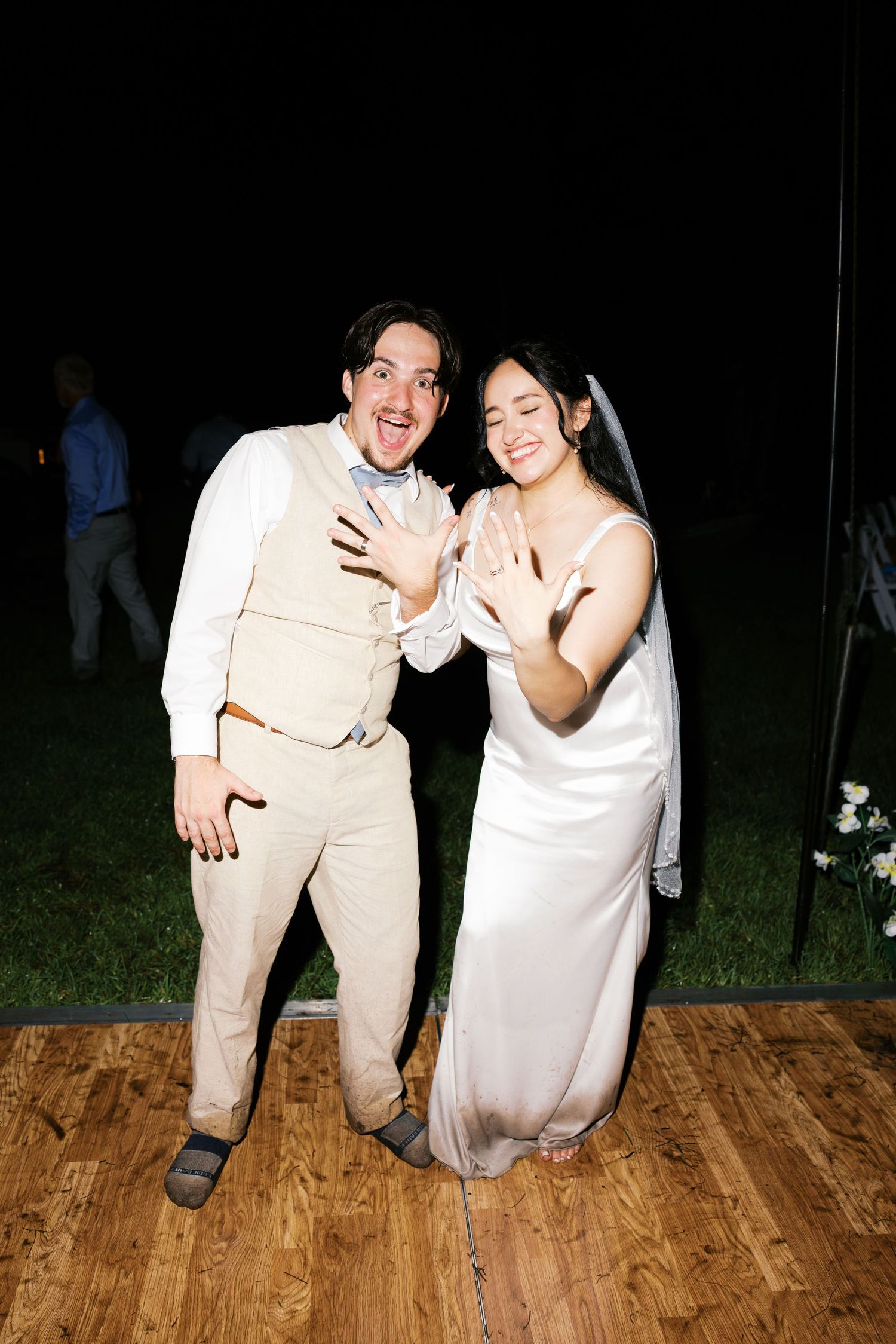 Newlywed couple joyfully shows off wedding rings on dance floor at outdoor nighttime wedding reception.