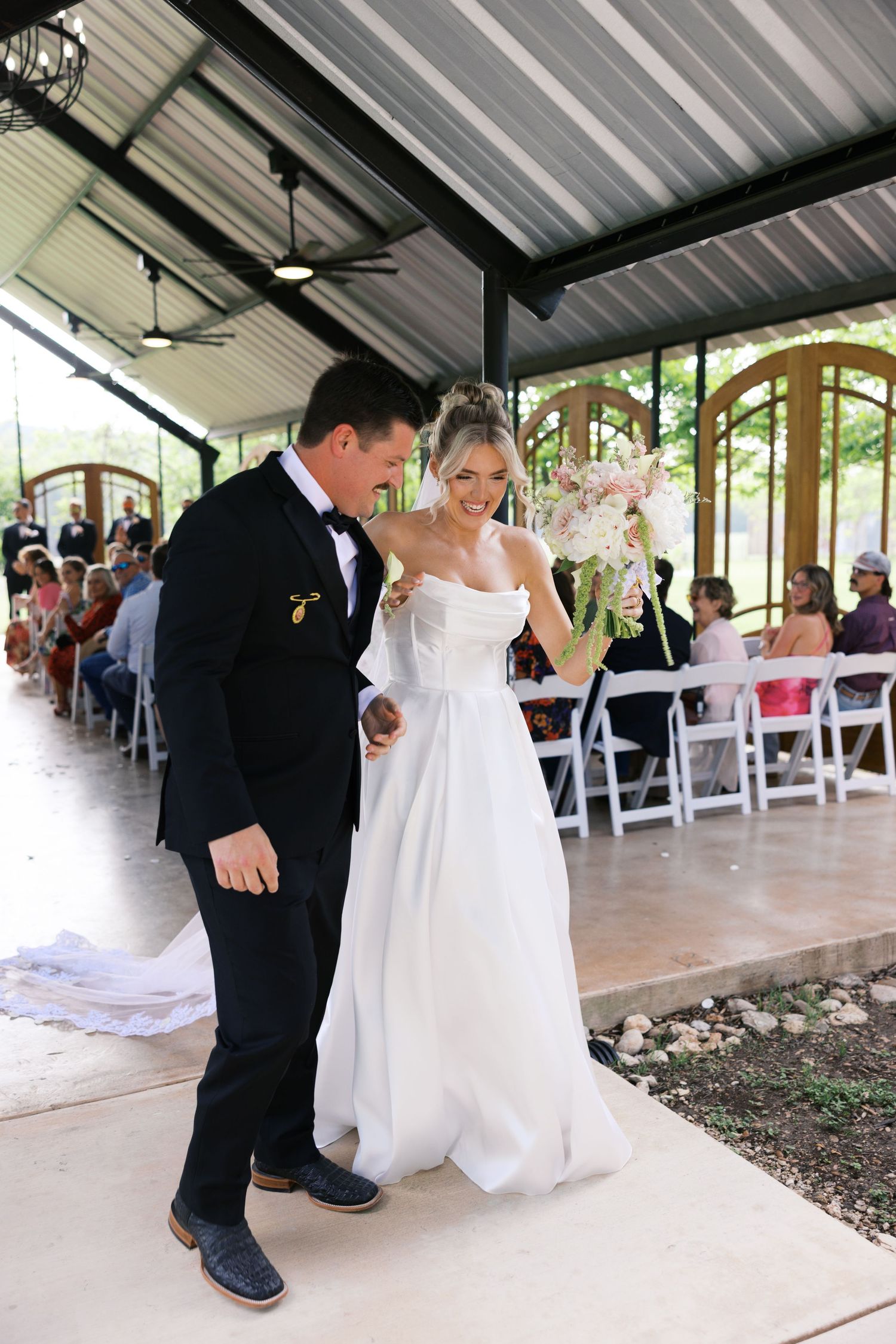 Bride and groom walk arm in arm down the aisle at an outdoor wedding ceremony under a covered pavilion.