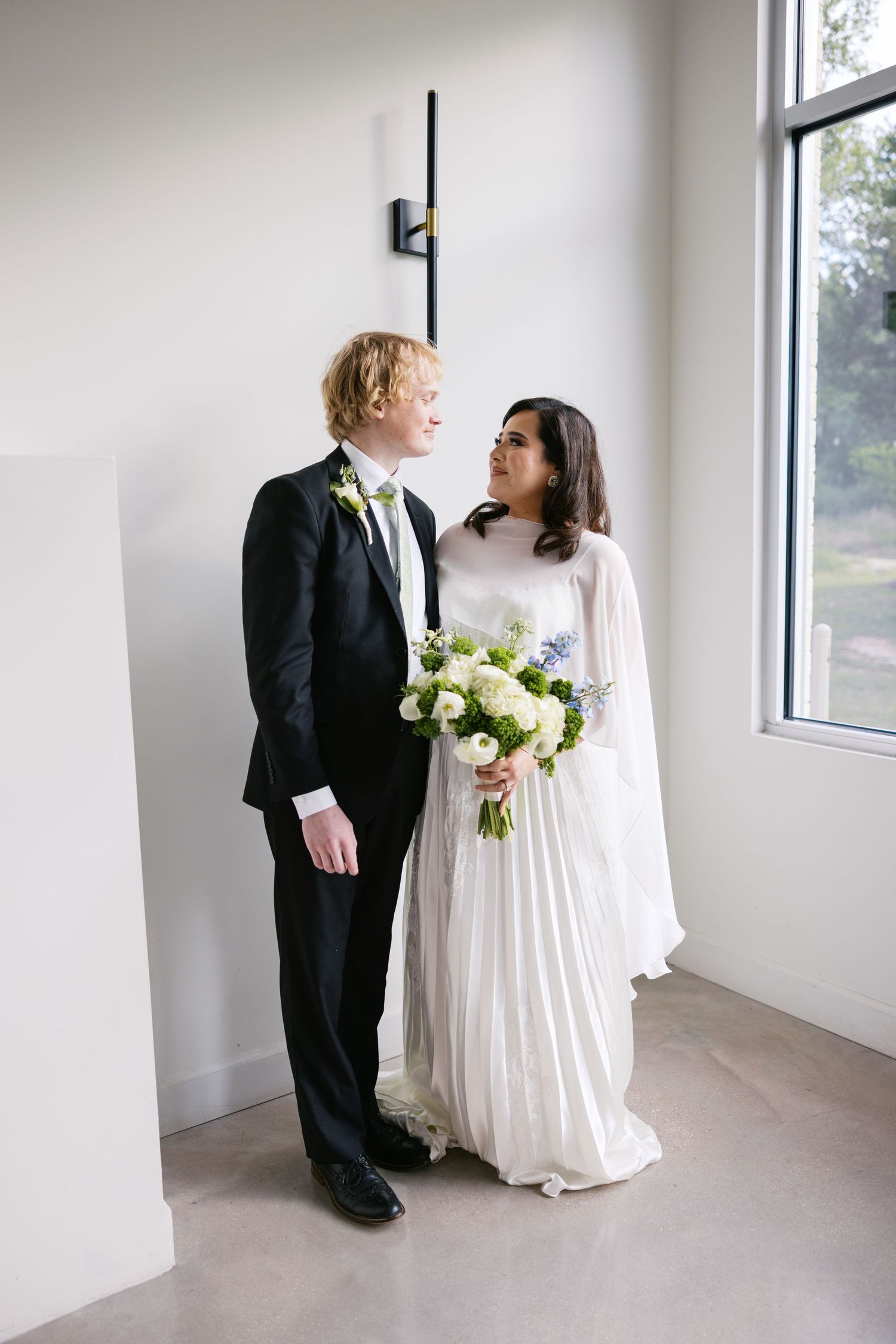 Bride and groom share a loving glance indoors, she holds a white bouquet, he wears a black tuxedo with green tie.