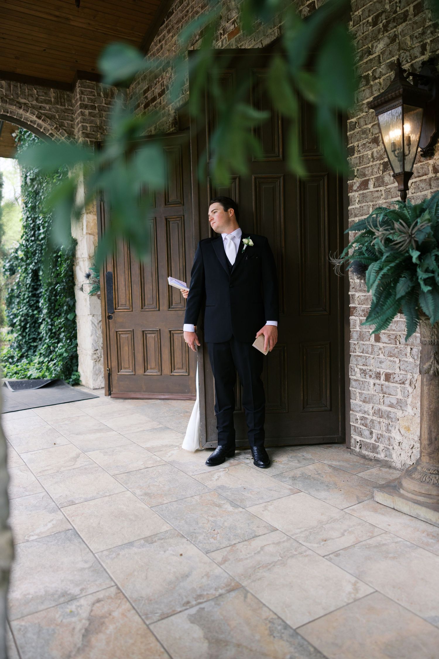 Groom in black tuxedo holding vows card waiting by ornate wooden doors with ivy-covered brick walls.