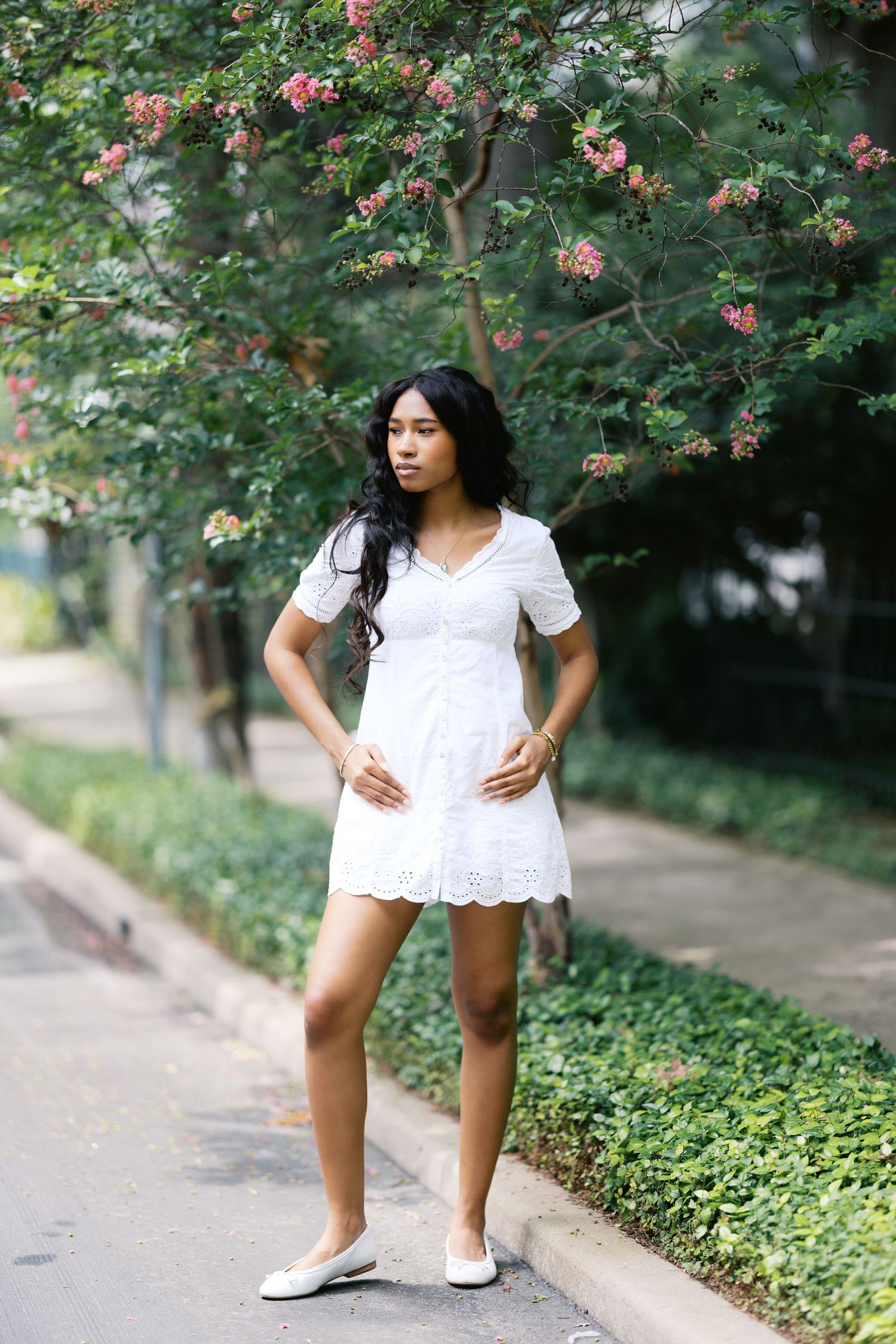 Young Black woman in white dress posing near pink flowering tree on garden path.