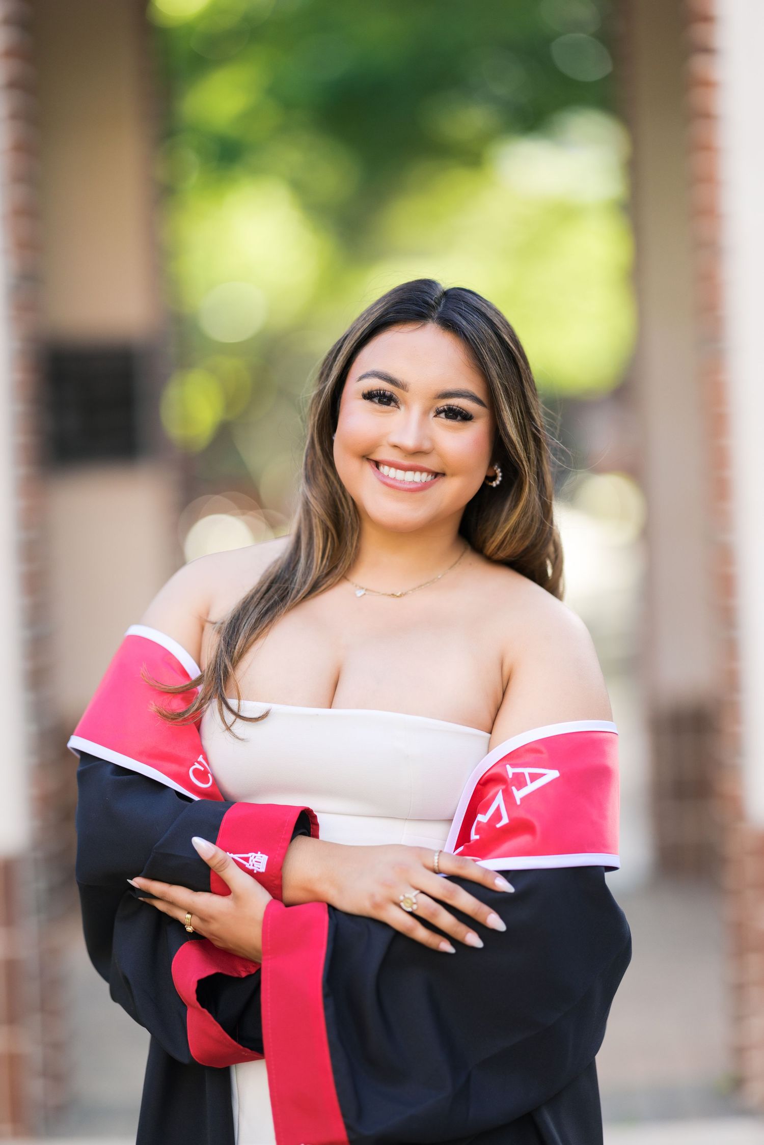 Smiling young woman in graduation gown with red trim posing outdoors for graduation photos.