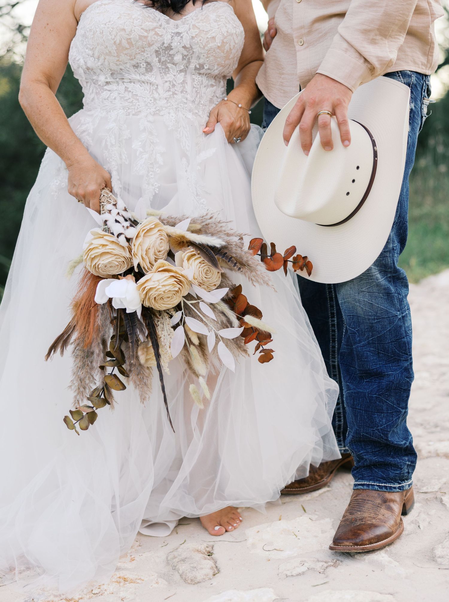 Close-up details of a rustic wedding outfit showing cowboy boots, white dress, and dried floral bouquet.