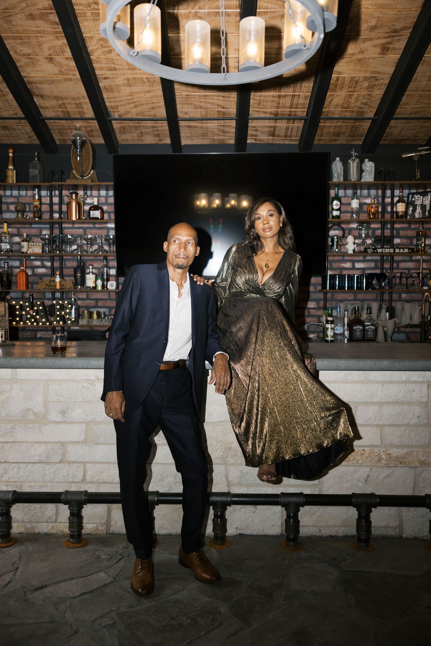 A couple poses at a rustic bar with exposed brick walls and wooden beams.