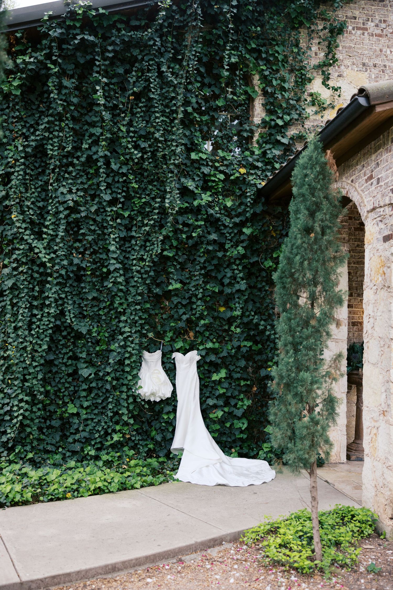 White wedding dress and flower girl dress hanging on ivy-covered brick wall at elegant outdoor venue.