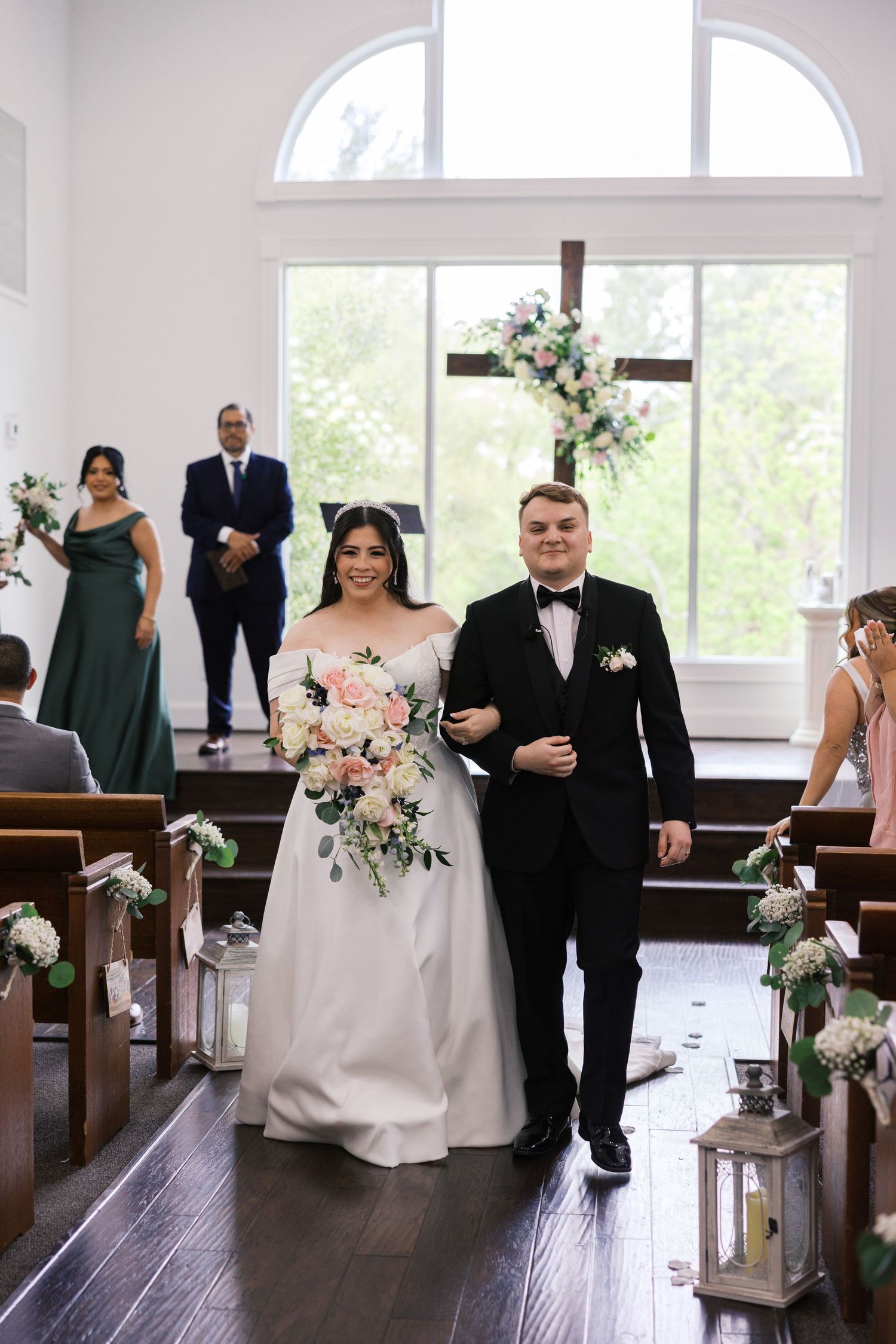 Bride and groom walk down the aisle of a white chapel after their wedding ceremony, surrounded by floral decorations.