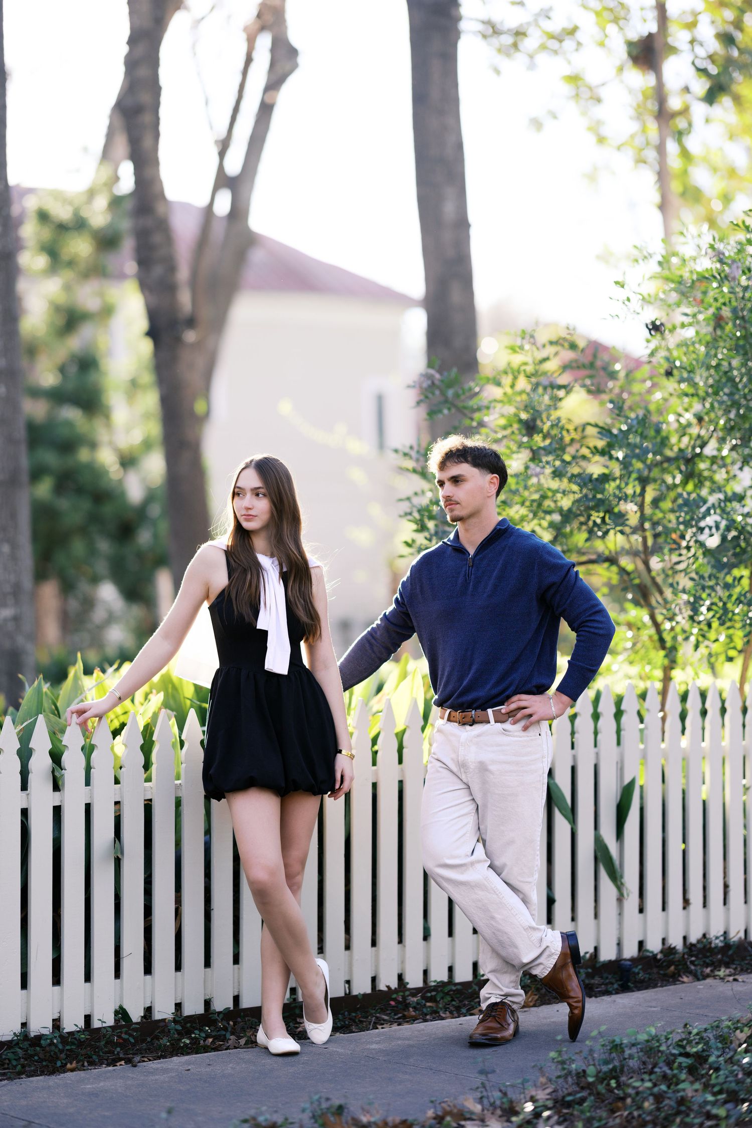 Young couple posing by a white picket fence outdoors, woman in black mini dress, man in navy blue sweater.