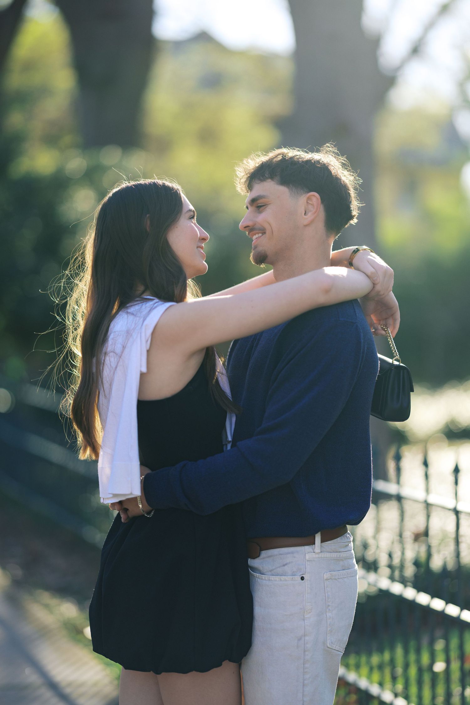 A young couple embraces and smiles at each other in a sunlit park, the woman in a black dress with a white jacket.