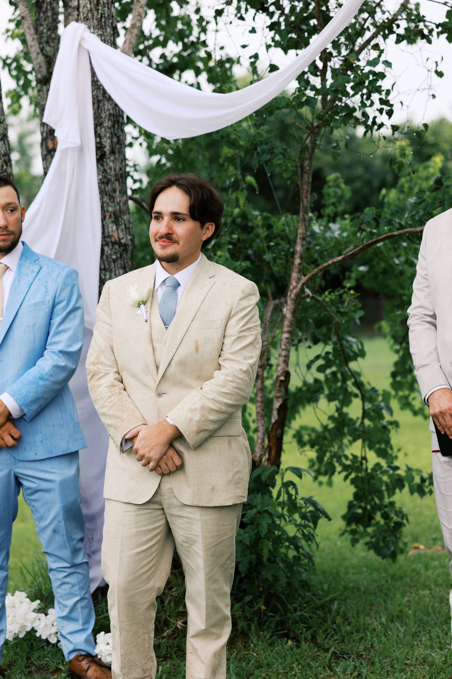Groom in beige suit and blue tie stands at outdoor wedding ceremony altar with groomsmen in light blue suits.