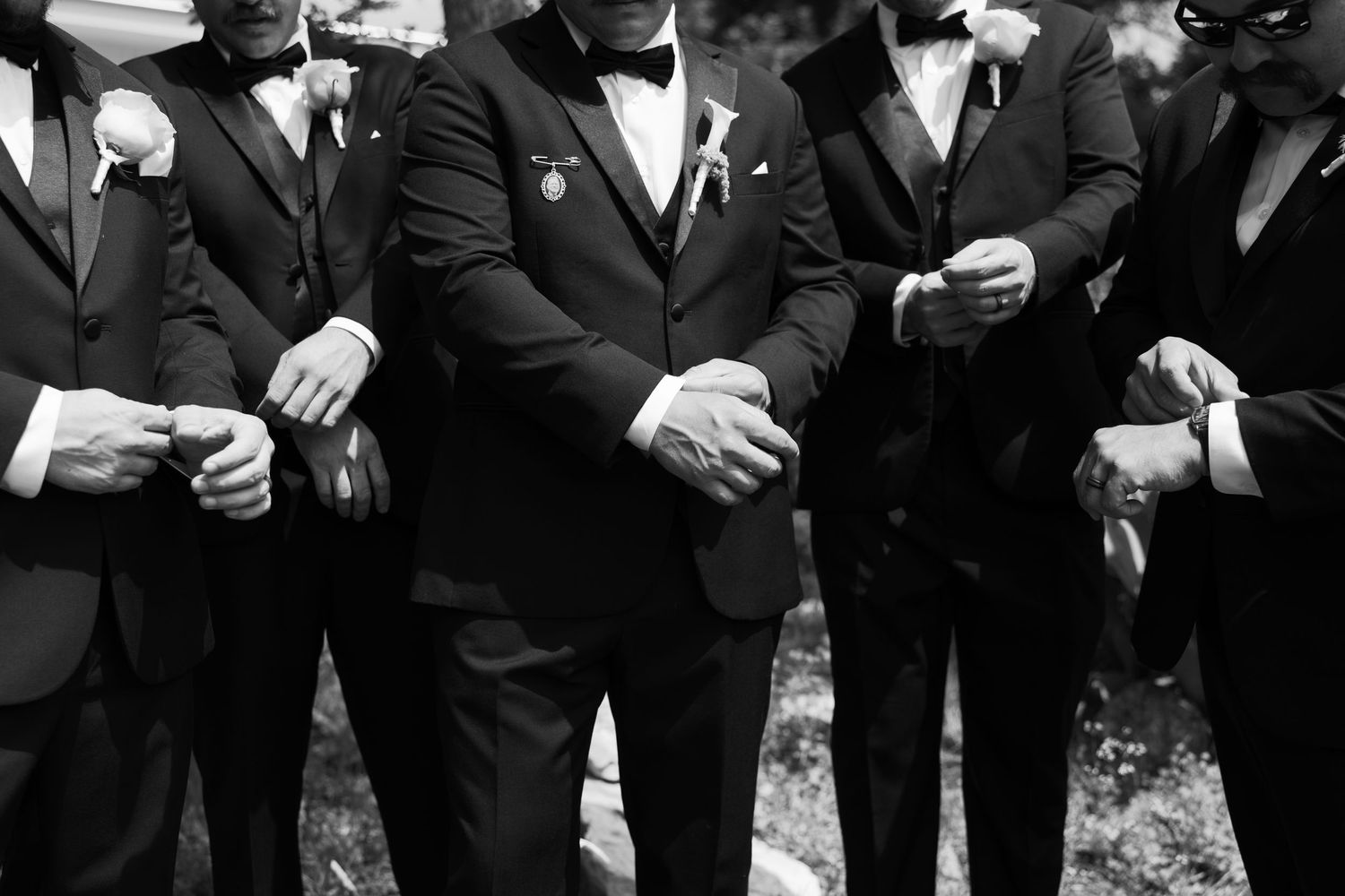 Black and white photo of groom and groomsmen in tuxedos adjusting cufflinks at a wedding.