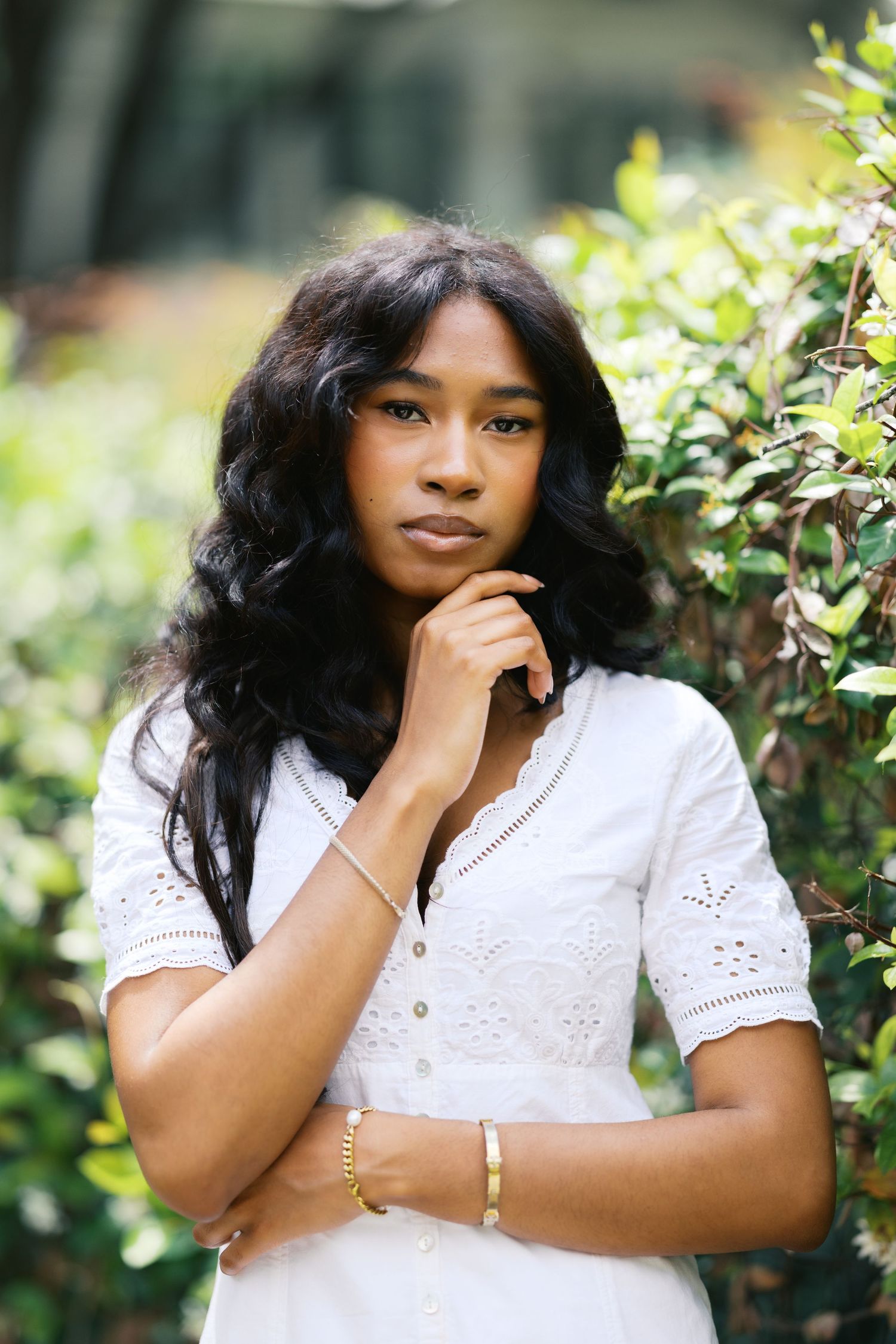 Young Black woman in white eyelet dress poses thoughtfully in lush green garden setting.