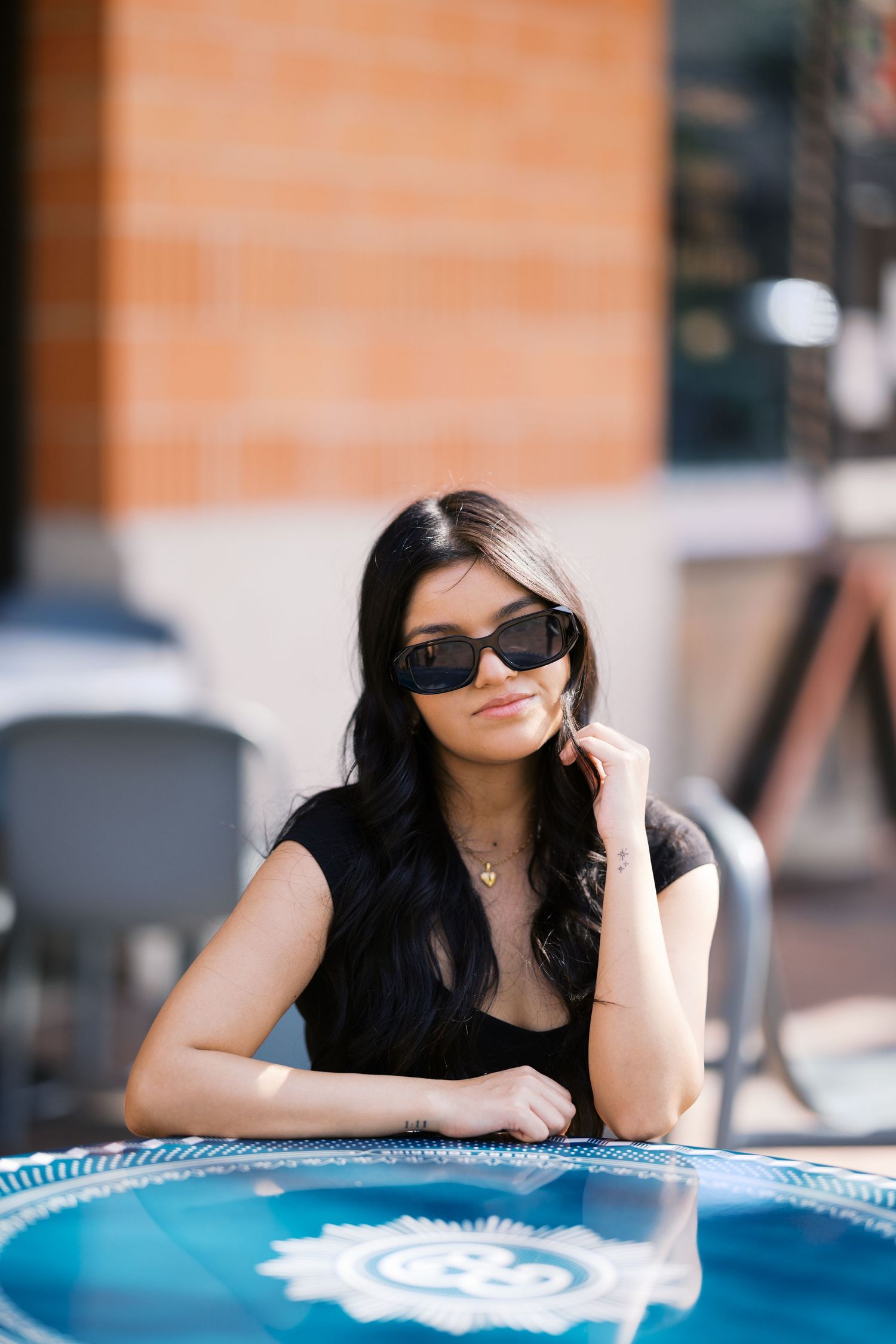 Relaxed pose at cafe table with blue umbrella showing summer style and sunglasses.