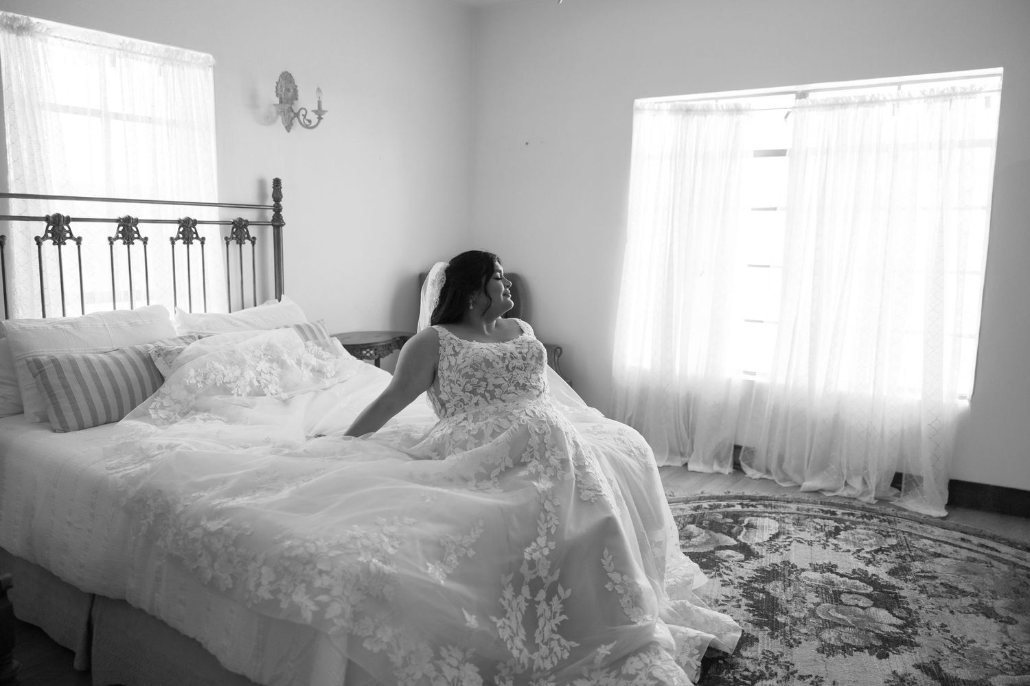A bride in a lace wedding dress sits on a bed near windows in a black and white photograph.