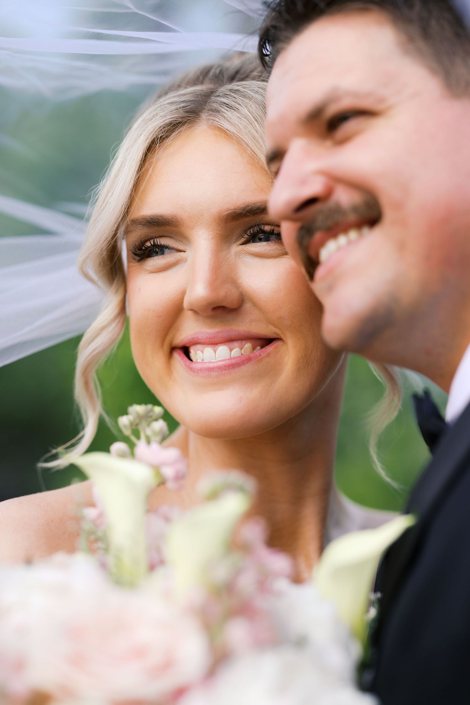 Smiling bride with veil and bouquet of white calla lilies poses closely with laughing groom outdoors.
