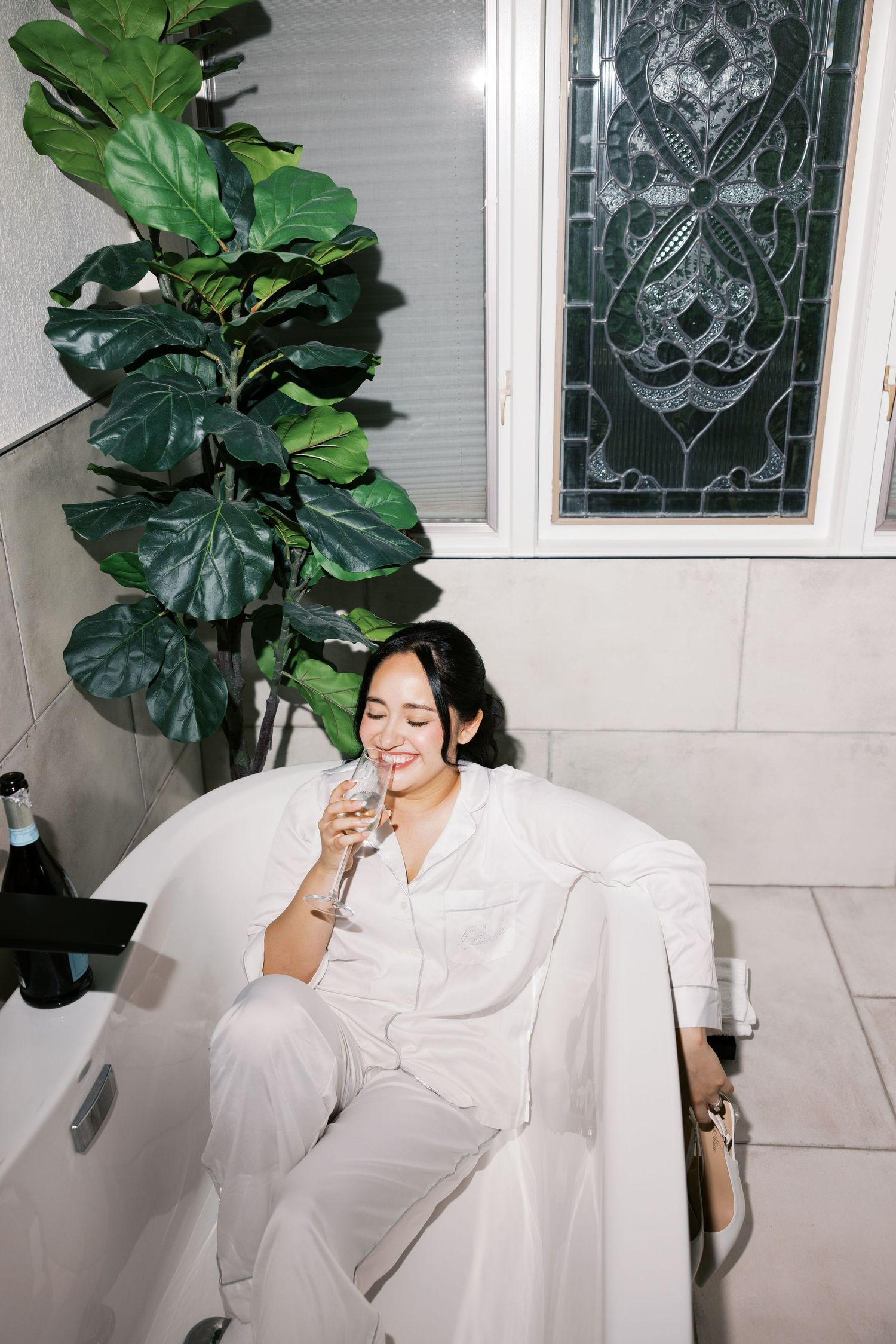 Woman in white outfit relaxing in bathtub with champagne glass, beside lush fiddle leaf fig plant.