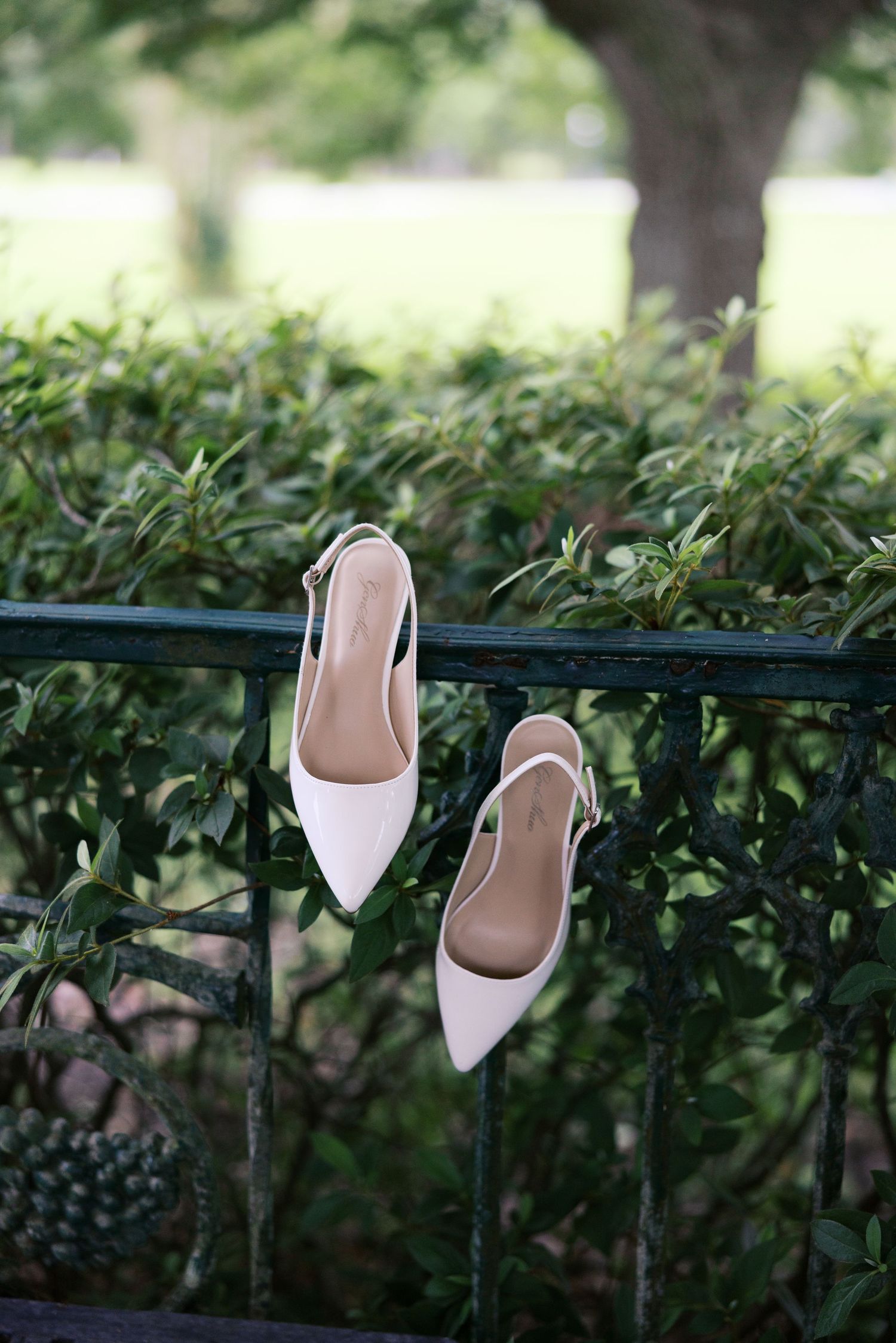 White slingback pointed-toe heels displayed on a black iron fence surrounded by lush green hedges outdoors.
