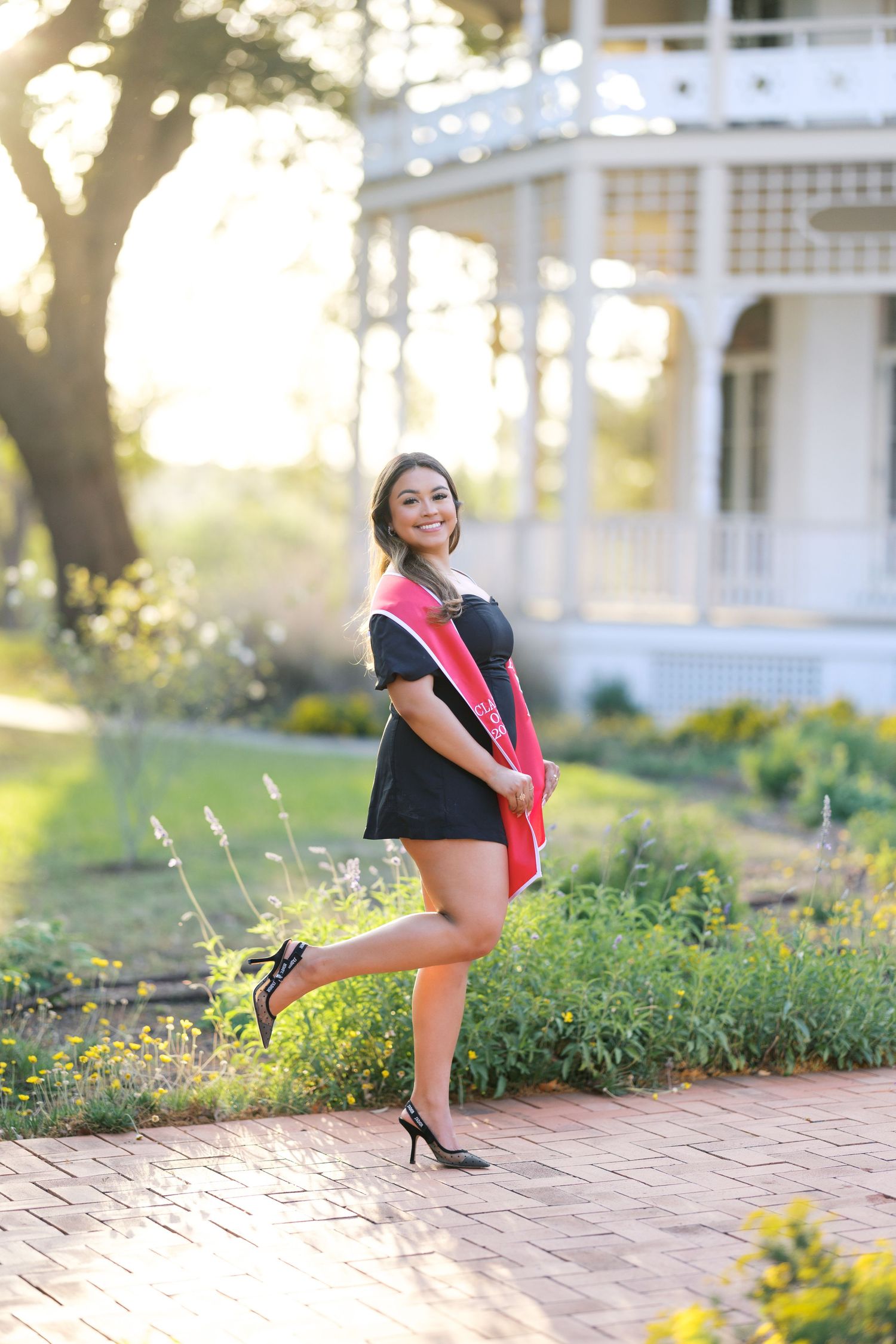 Smiling graduate in black dress with red sash poses playfully on brick path near white Victorian building.