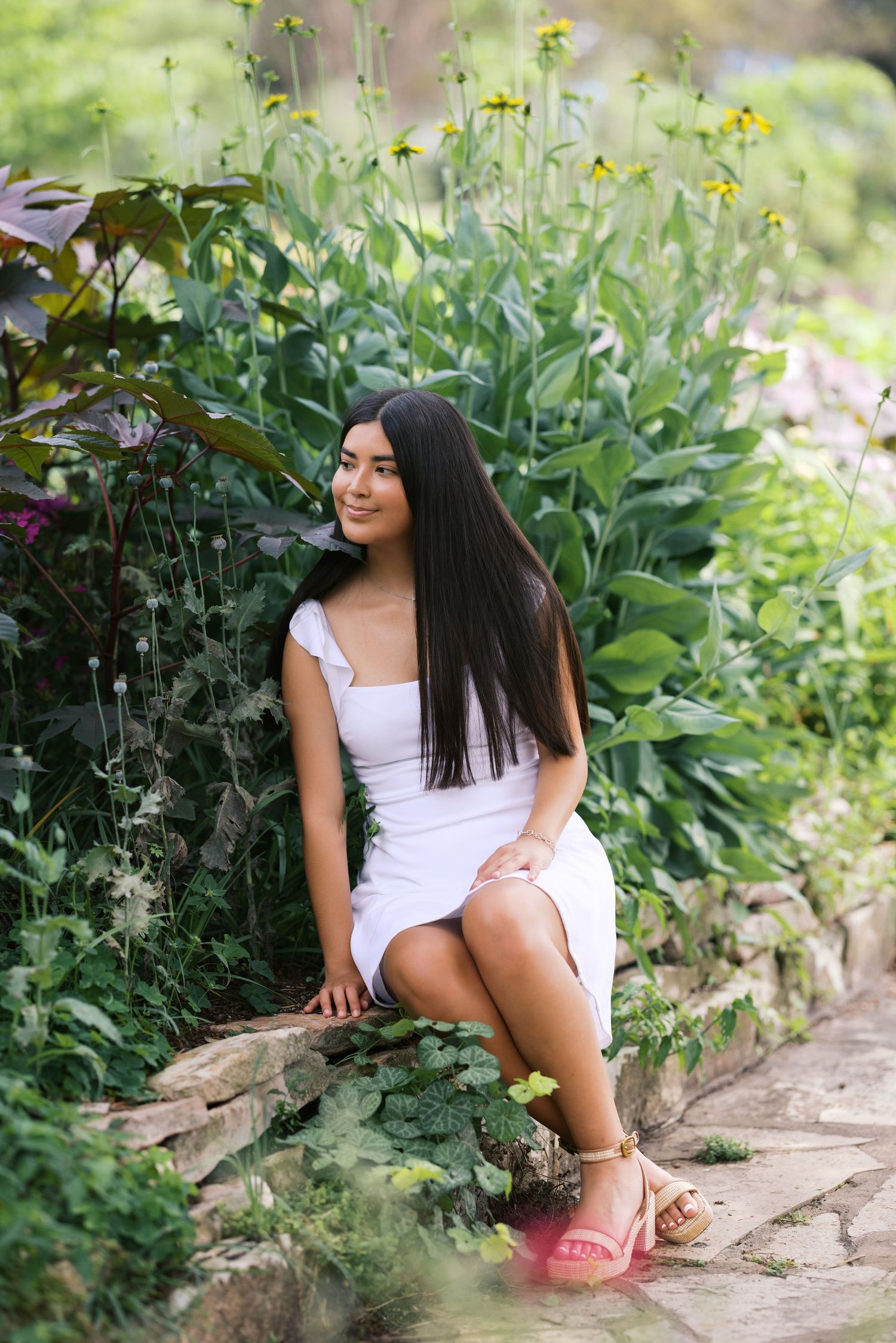 Young woman in white dress sitting on stone wall surrounded by vibrant garden flowers.