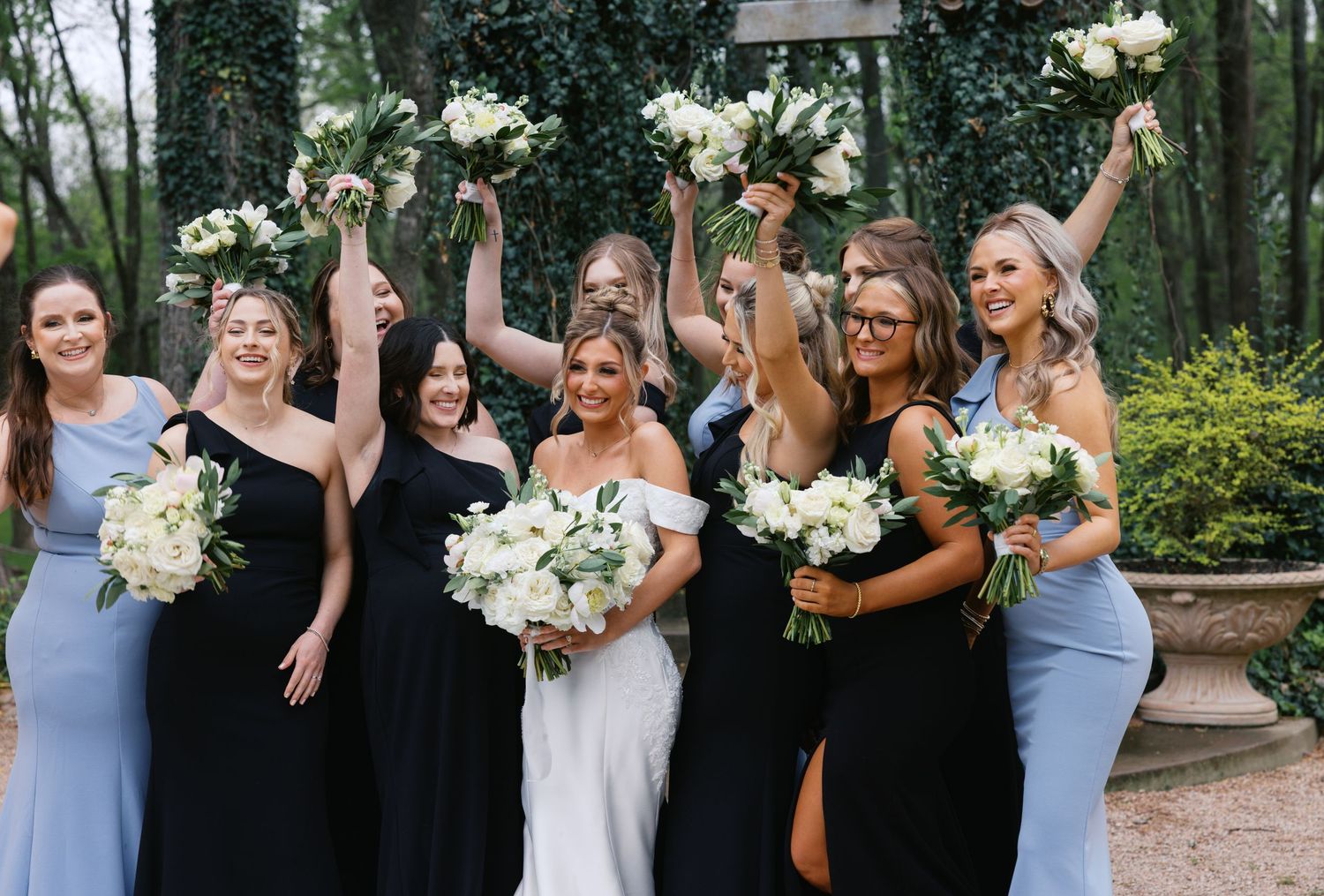 Bride in white gown celebrates with bridesmaids in blue and black dresses, holding white floral bouquets outdoors.
