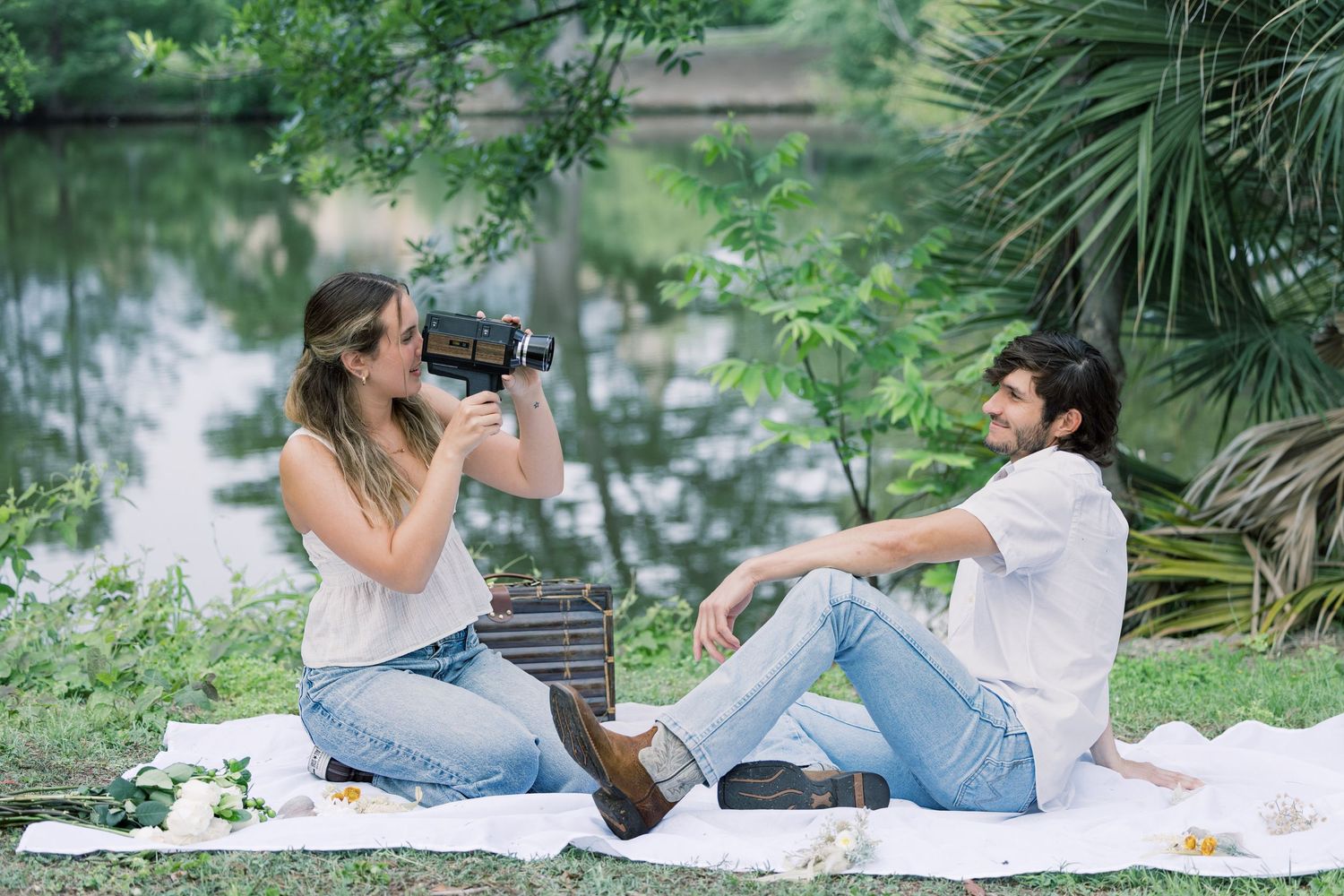 A couple lounges on a picnic blanket surrounded by lush greenery near water.