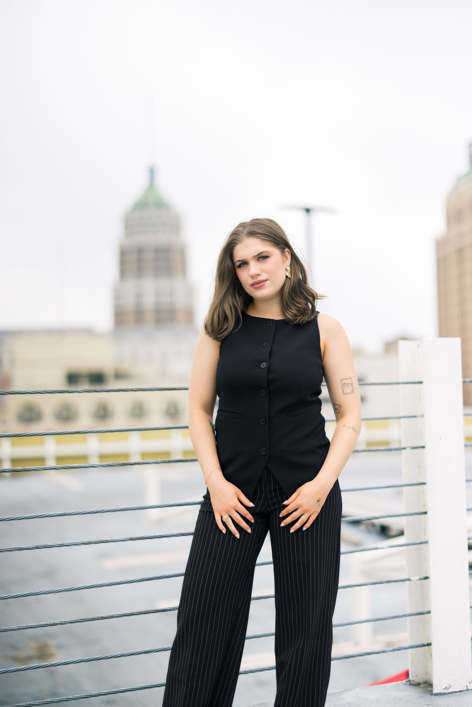 Portrait of person in black vest and pinstripe pants on rooftop with city buildings in background.