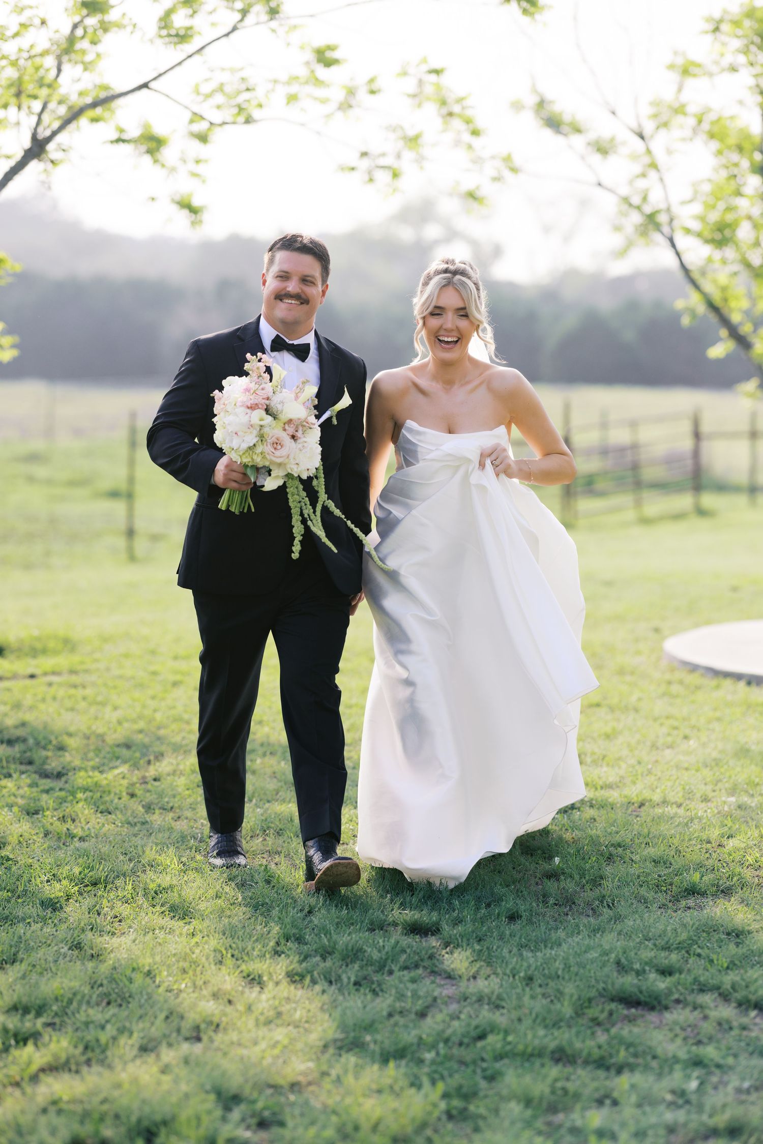 Joyful bride and groom with mustache walk through green field, groom holding white floral bouquet.