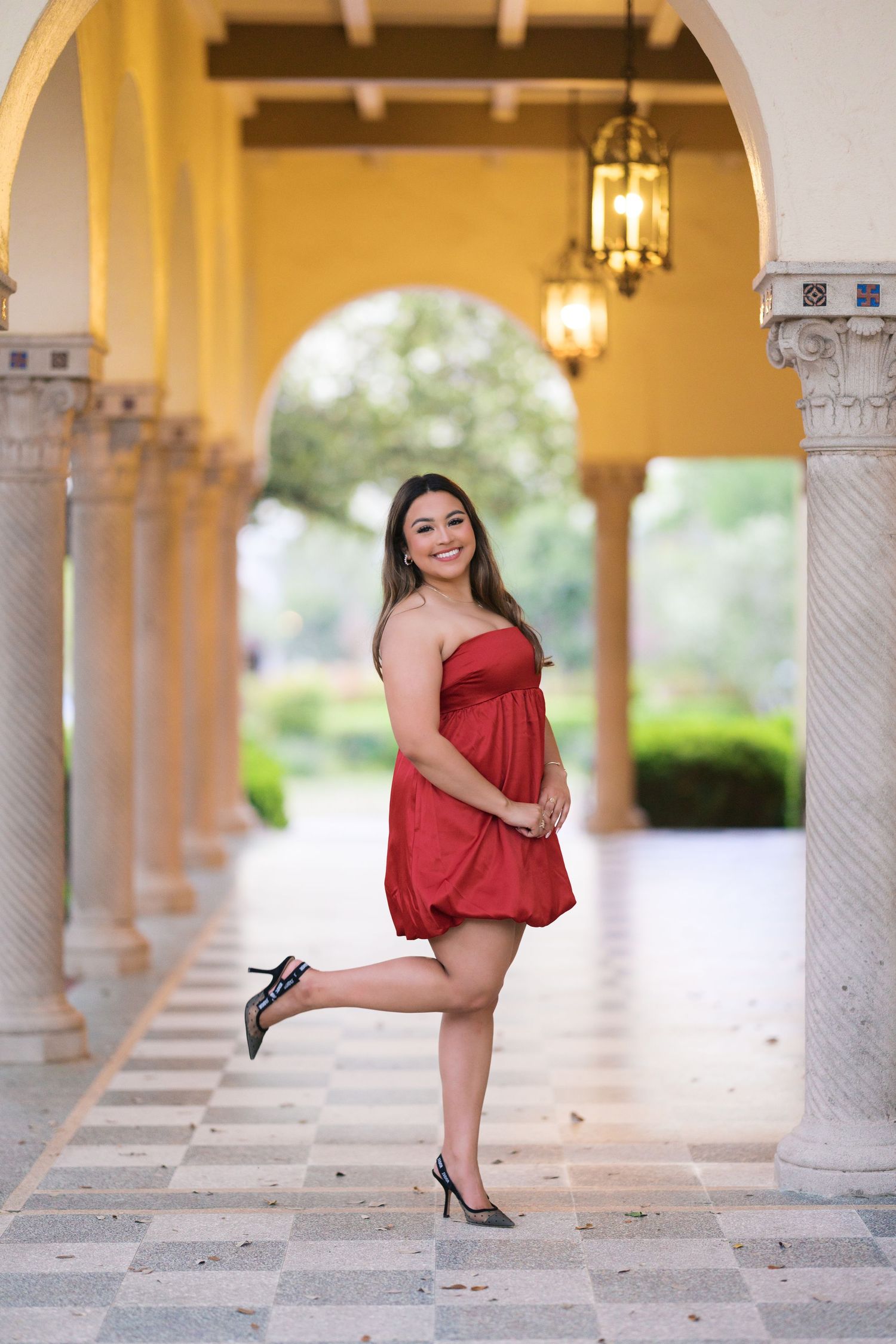 Young woman in red strapless mini dress posing on one leg in elegant arched colonnade with hanging lanterns.