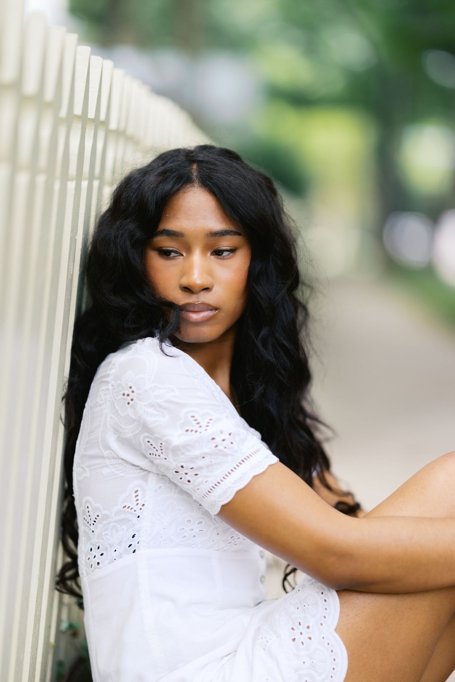 Young woman with long wavy hair in white eyelet dress sits pensively against white picket fence outdoors.