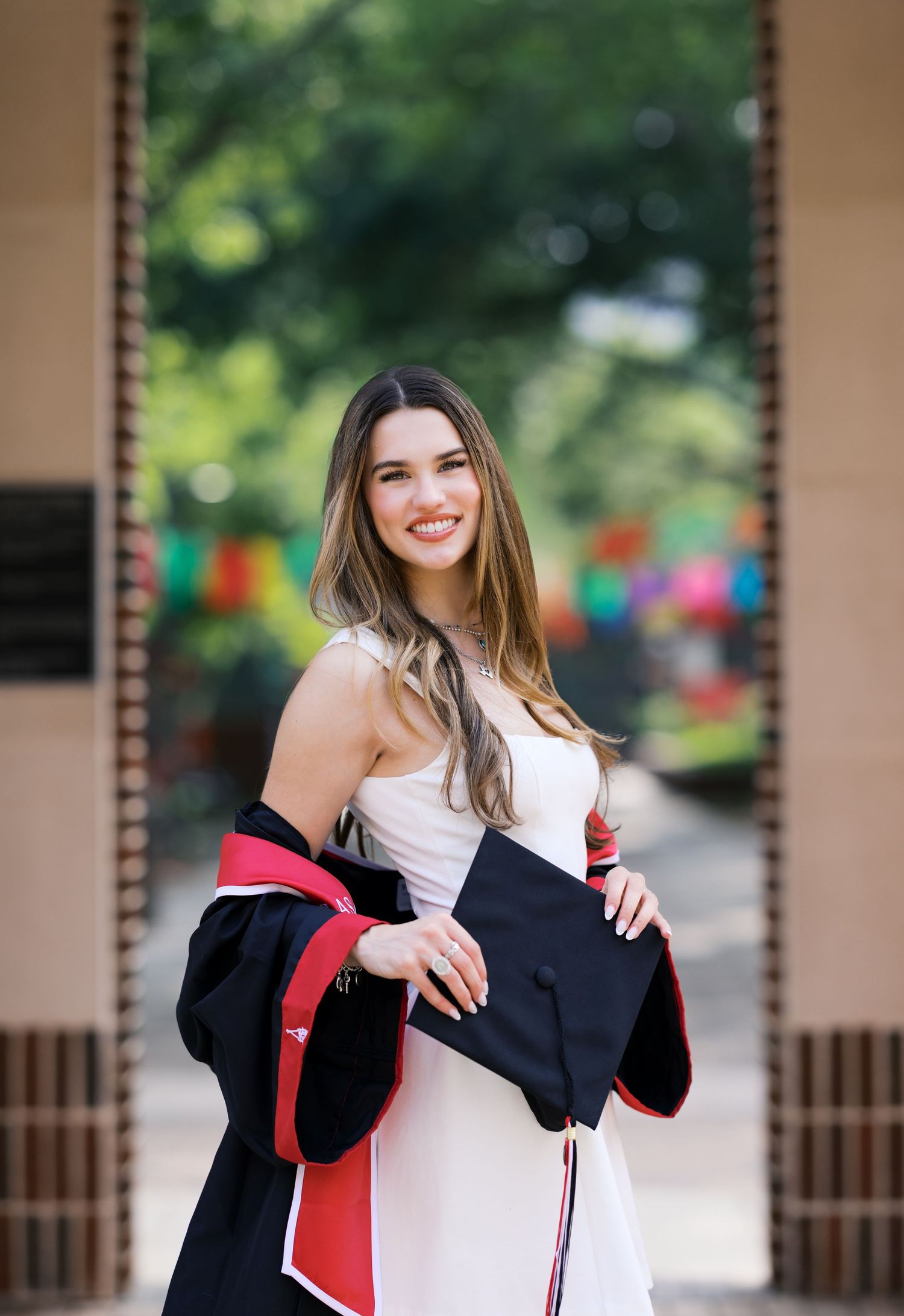 Smiling female graduate in white dress holding black and red graduation gown and cap outdoors.