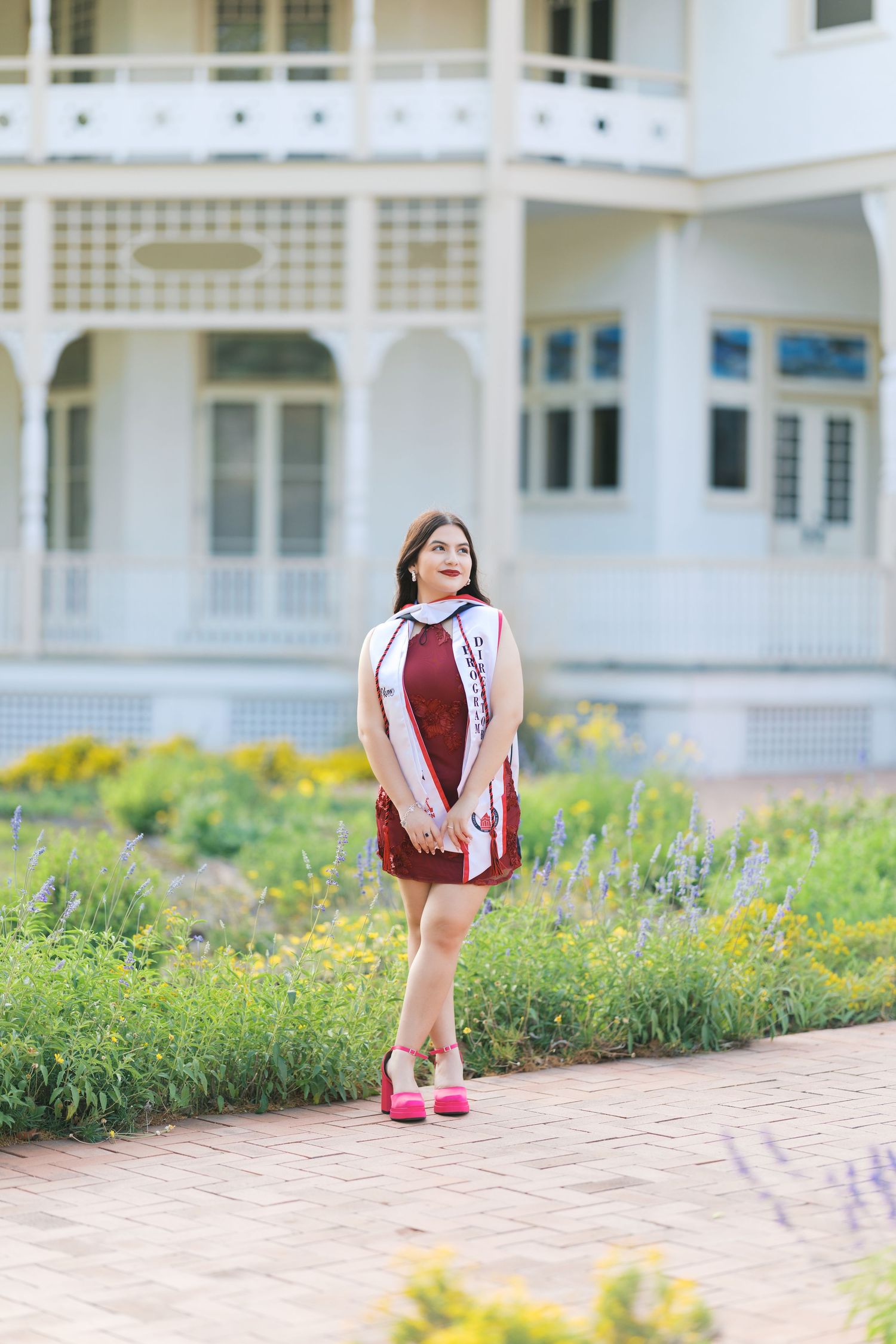 Young woman in maroon dress and sash posing on brick path before a white Victorian building with gardens.