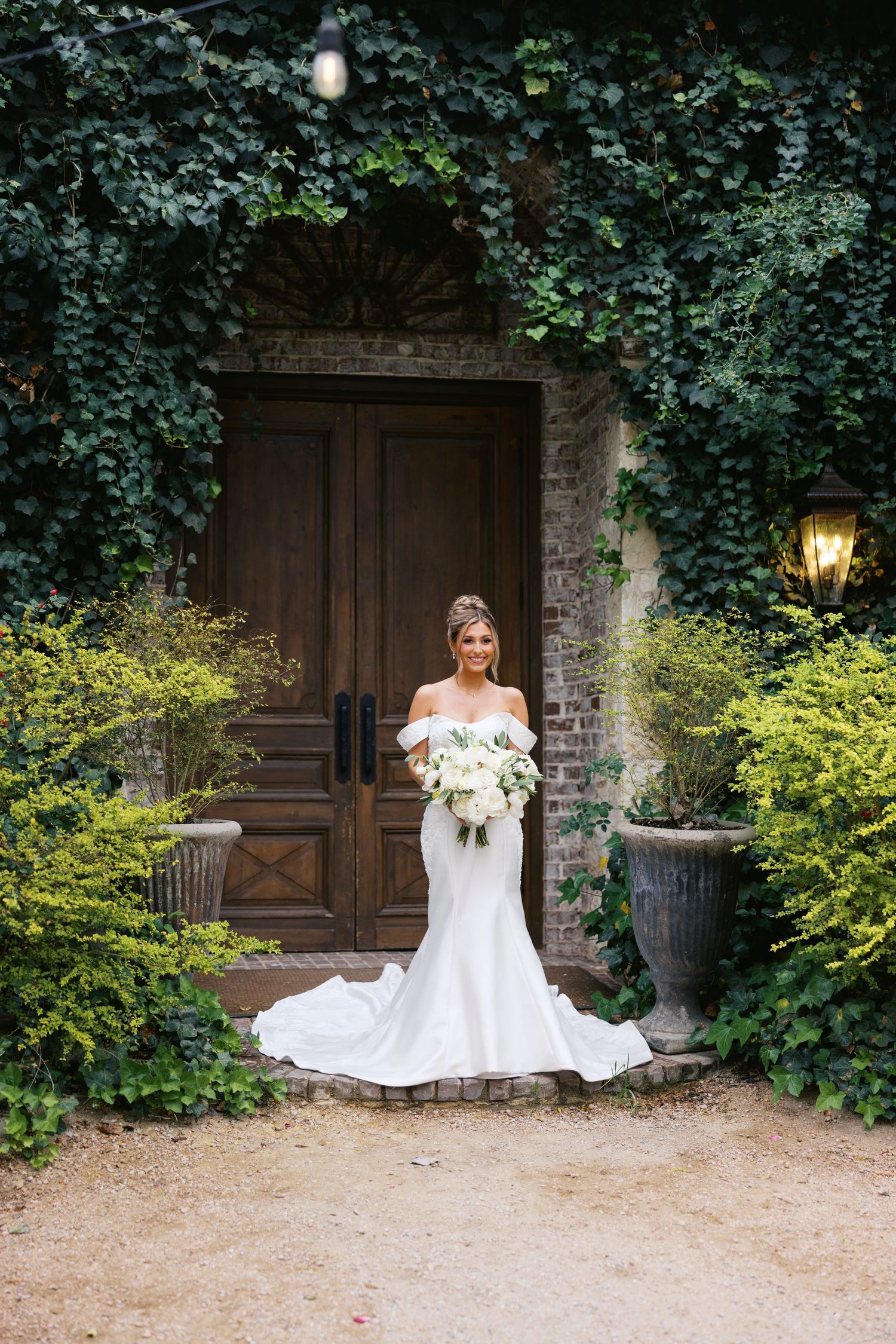 Bride in off-shoulder white mermaid wedding gown holding bouquet before ivy-covered brick building with wooden doors.