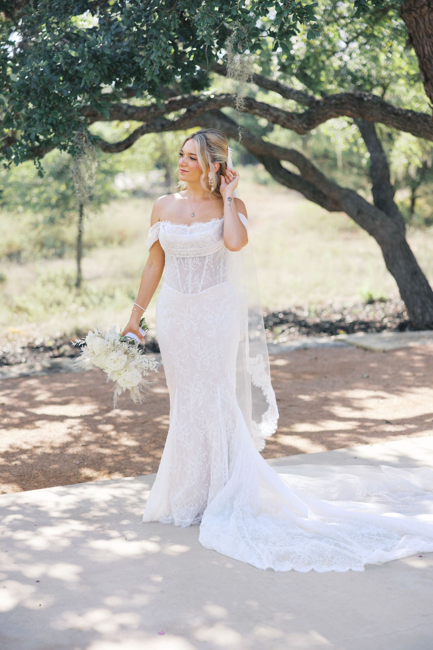 Bride in fitted lace wedding dress with off-shoulder detail poses in dappled sunlight.
