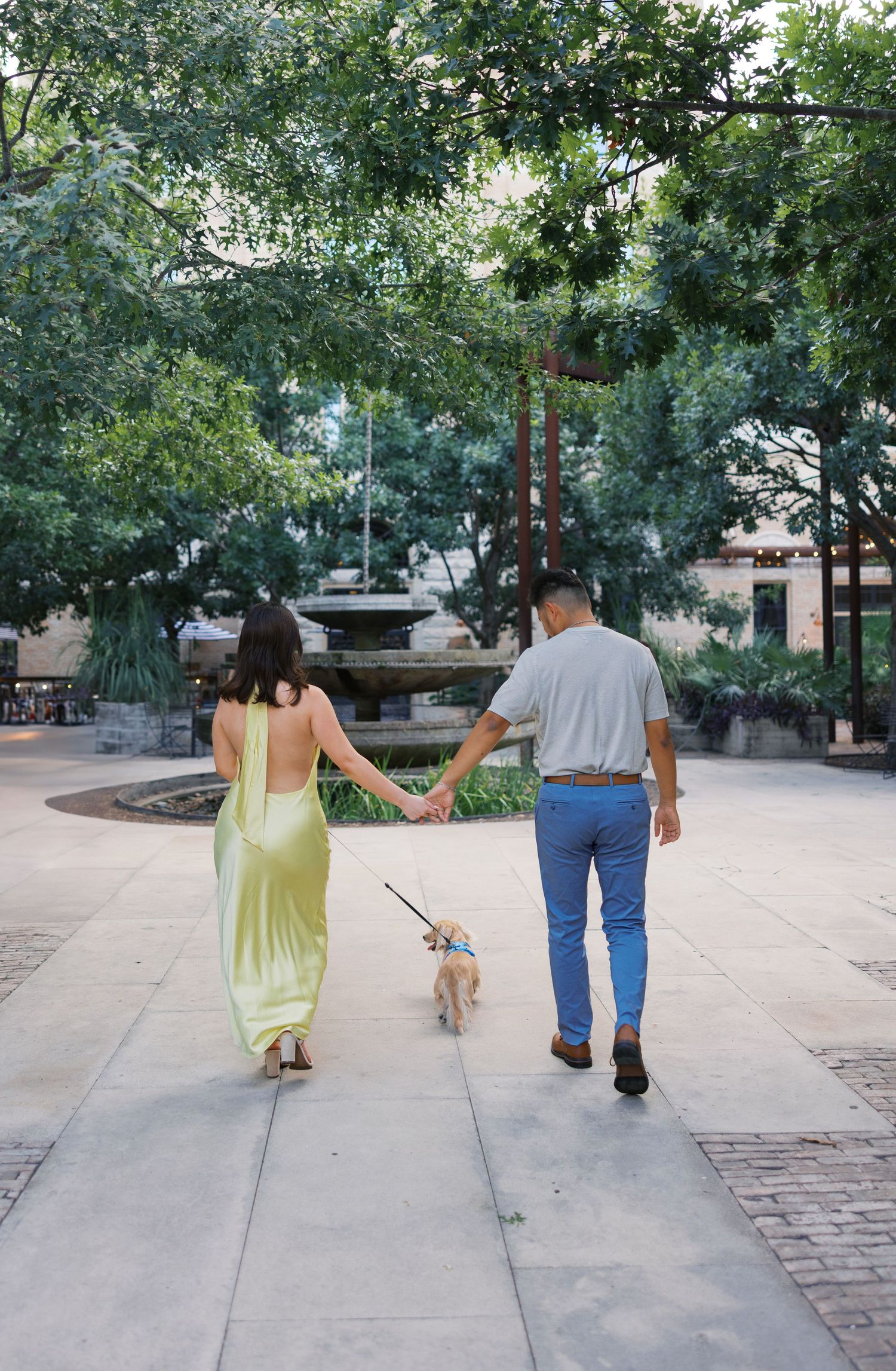 Couple walking hand in hand on brick pathway under tree canopy.